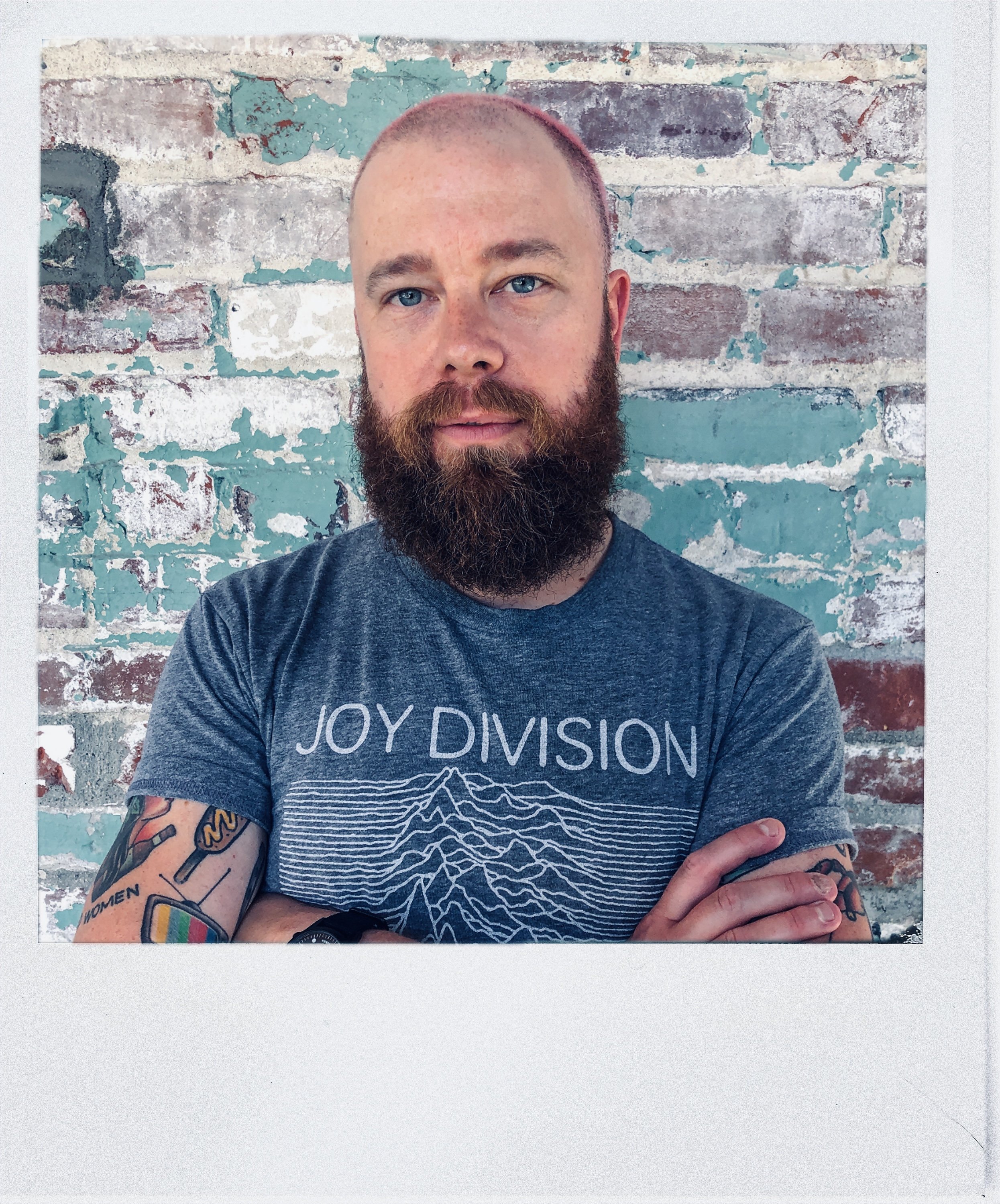 A man with a beard and short hair wearing a Joy Division t-shirt, standing with his arms crossed in front of a weathered brick wall with teal and red paint peeling.