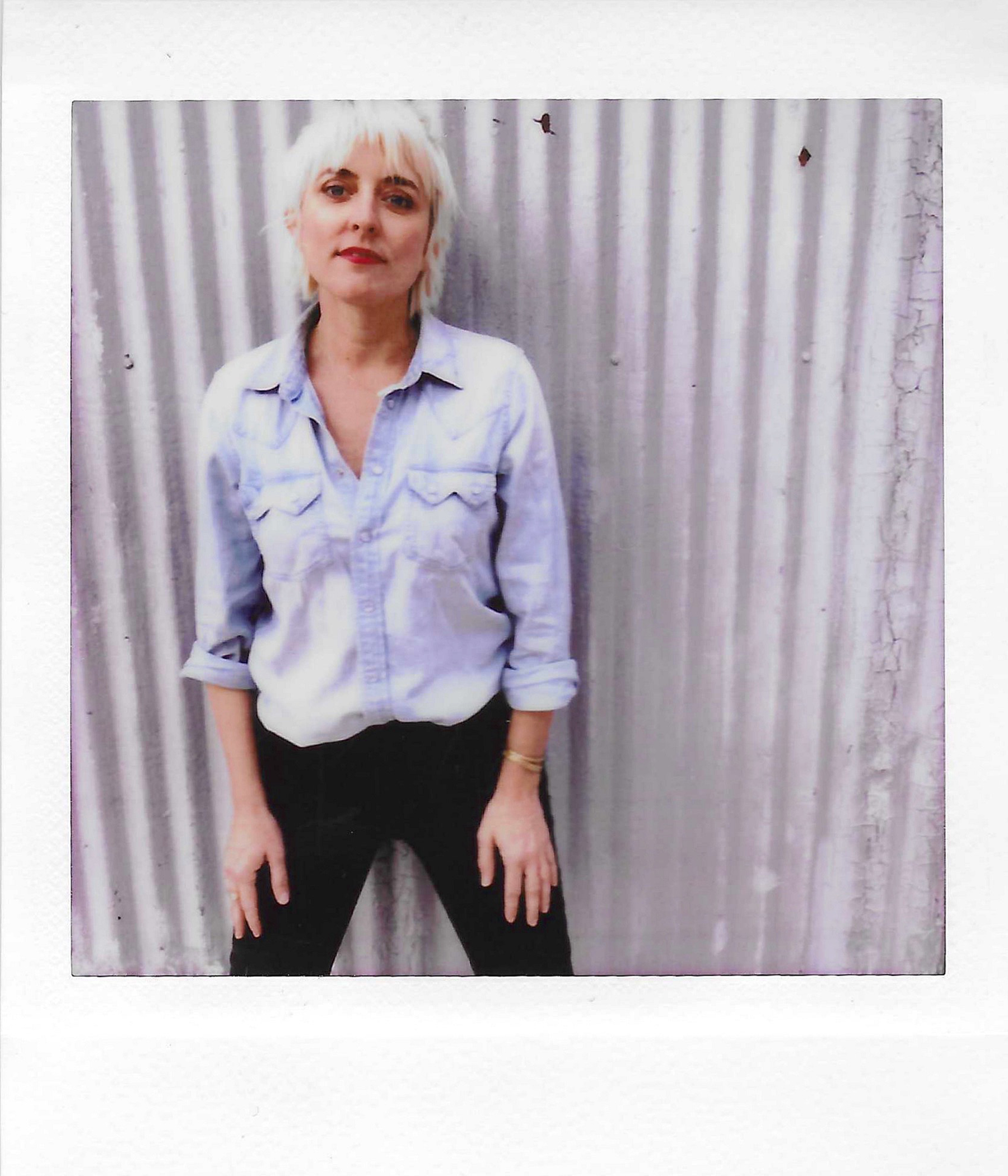 Photograph of a woman with short blonde hair, wearing a light blue button-up shirt, standing in front of a corrugated metal background, with her hands resting on her thighs.