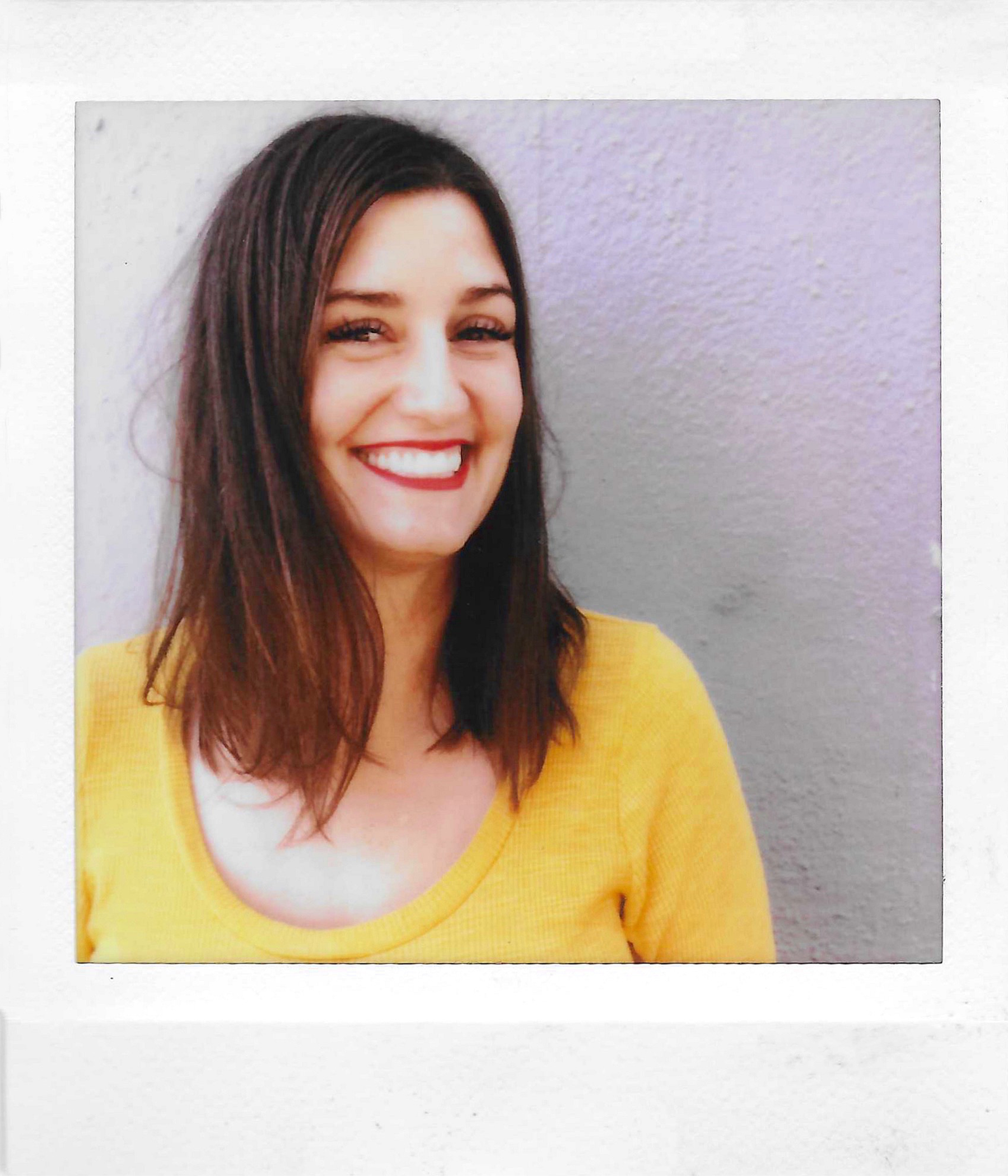Close-up of a smiling woman with long brown hair, wearing a yellow top, standing in front of a gray wall.