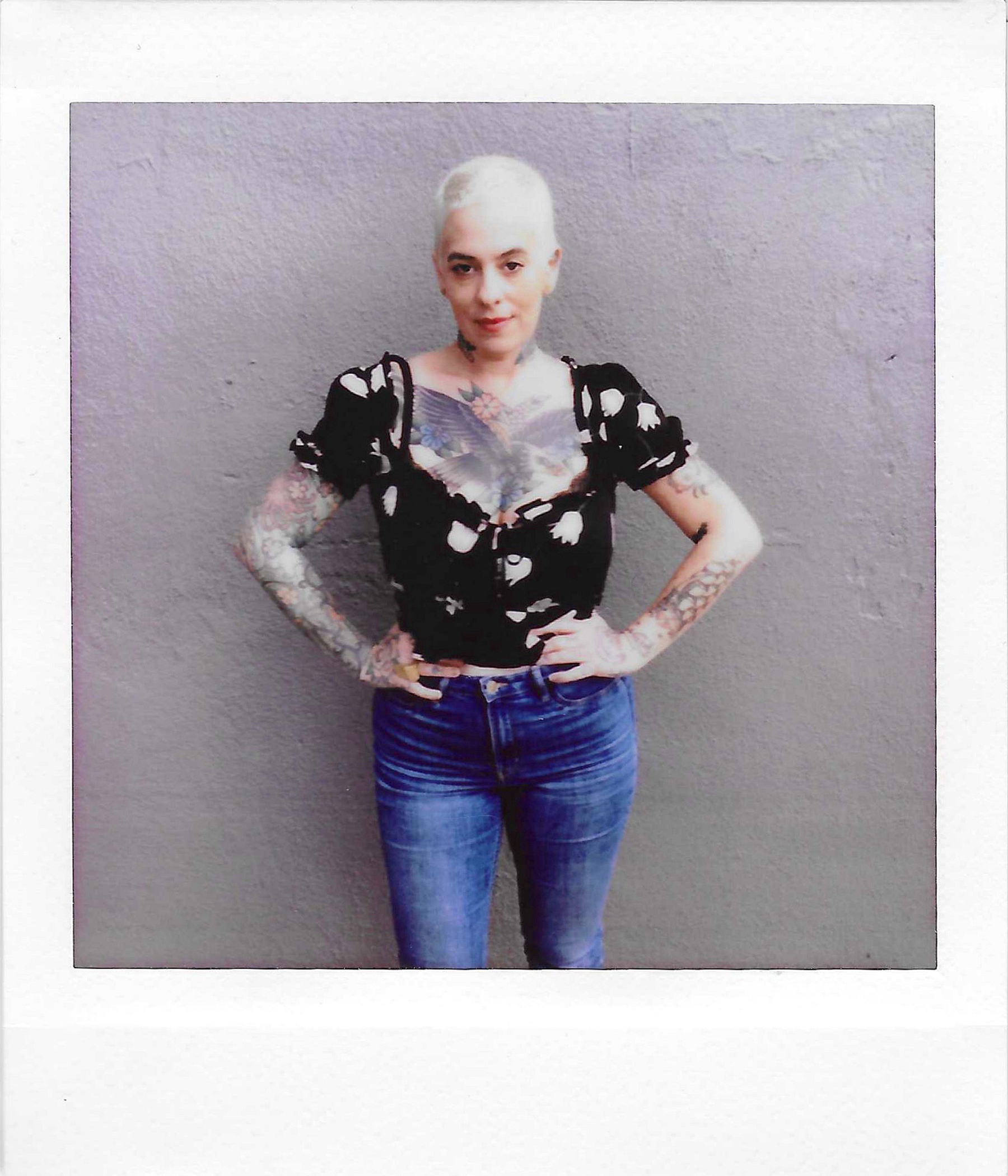 A woman with platinum blonde hair, tattoos on her arms and chest, wearing a black blouse with white puffed sleeves and a floral pattern, paired with blue jeans, stands against a gray textured wall.