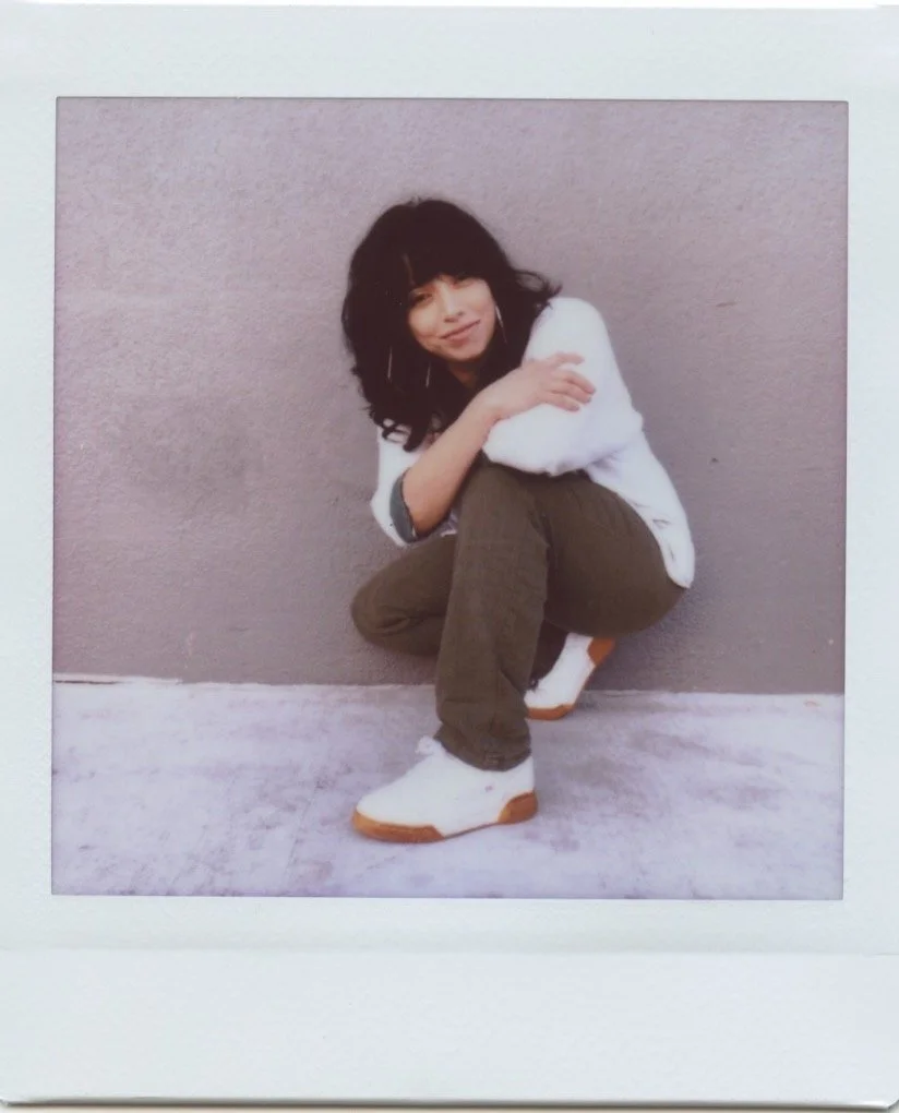 A young woman with black hair, wearing a white sweater, brown pants, and white sneakers, crouching against a gray wall, smiling at the camera.
