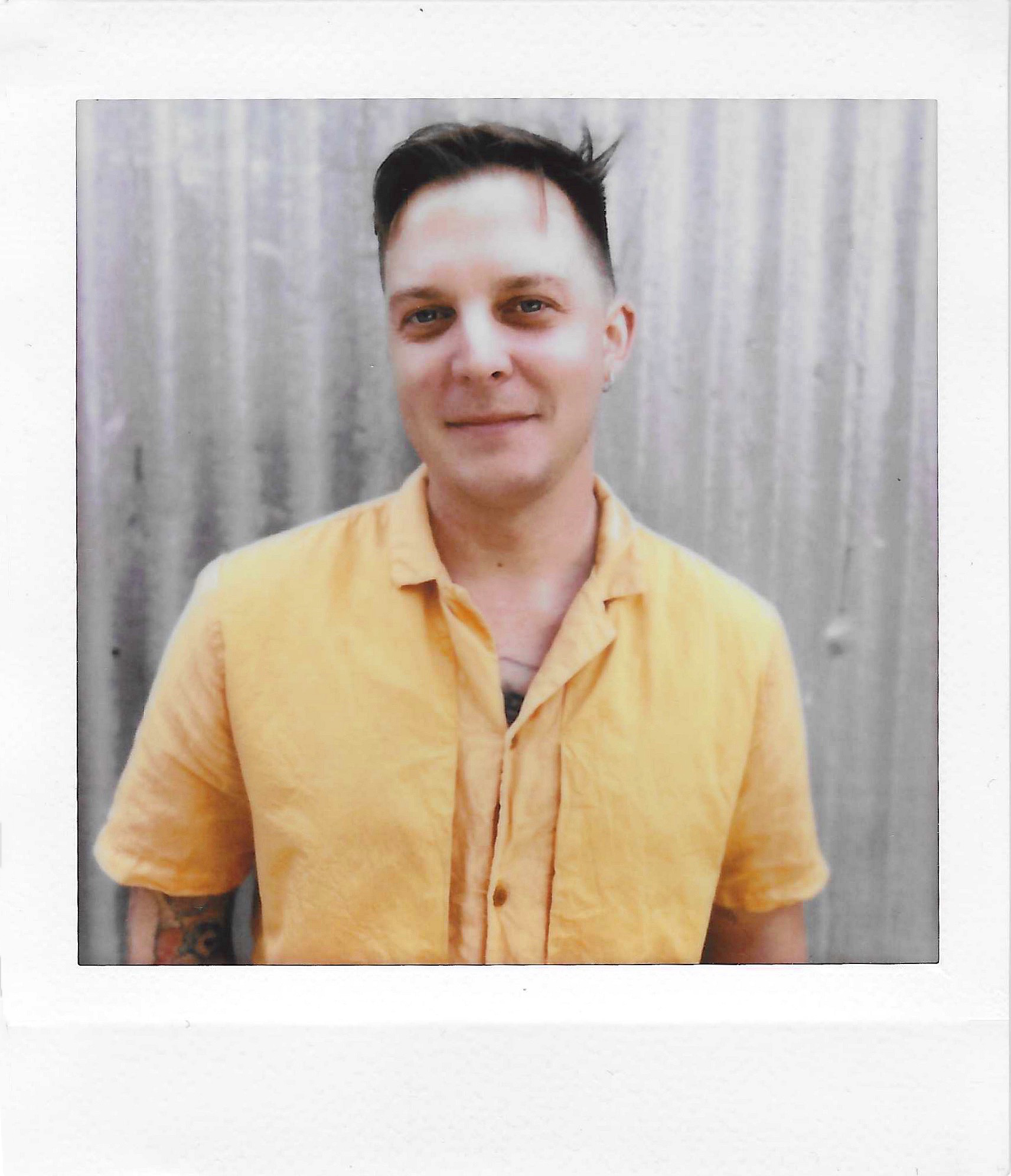 A young person with short dark hair and light skin tone wearing a yellow button-up shirt, standing in front of a plain, light-colored, textured curtain background.