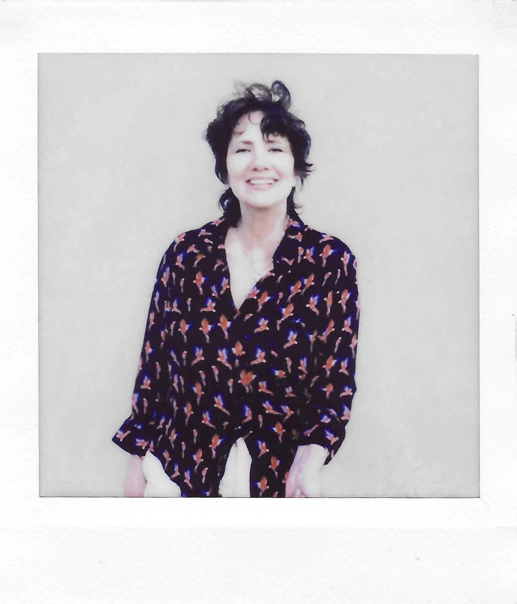 A woman with short dark curly hair smiling, wearing a dark floral blouse, standing against a plain light-colored background.