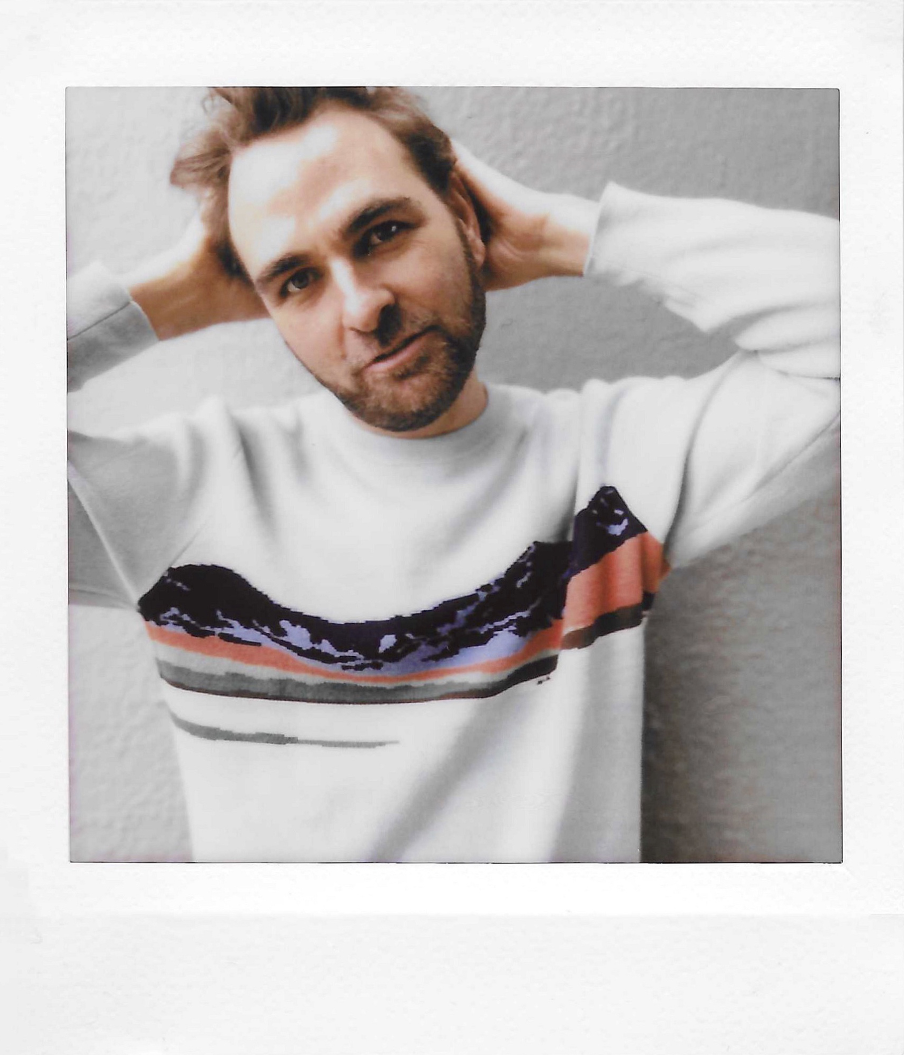 A man with brown hair and a beard posing with his hands behind his head, wearing a white sweater with a colorful pattern on the front, against a plain gray background.