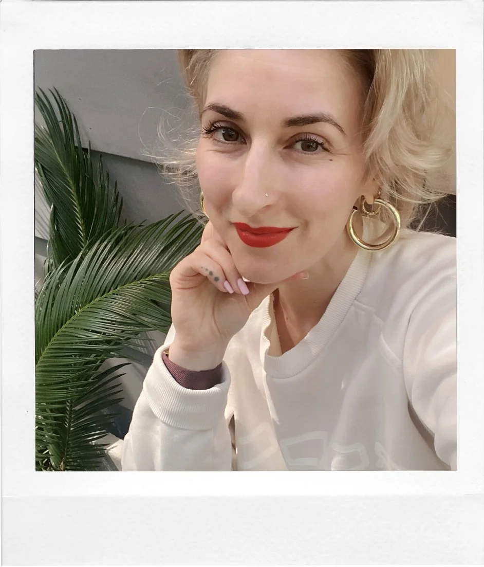 A woman with blonde, curly hair and red lipstick smiling and resting her chin on her hand, wearing gold hoop earrings and a cream-colored top, with green palm leaves behind her.