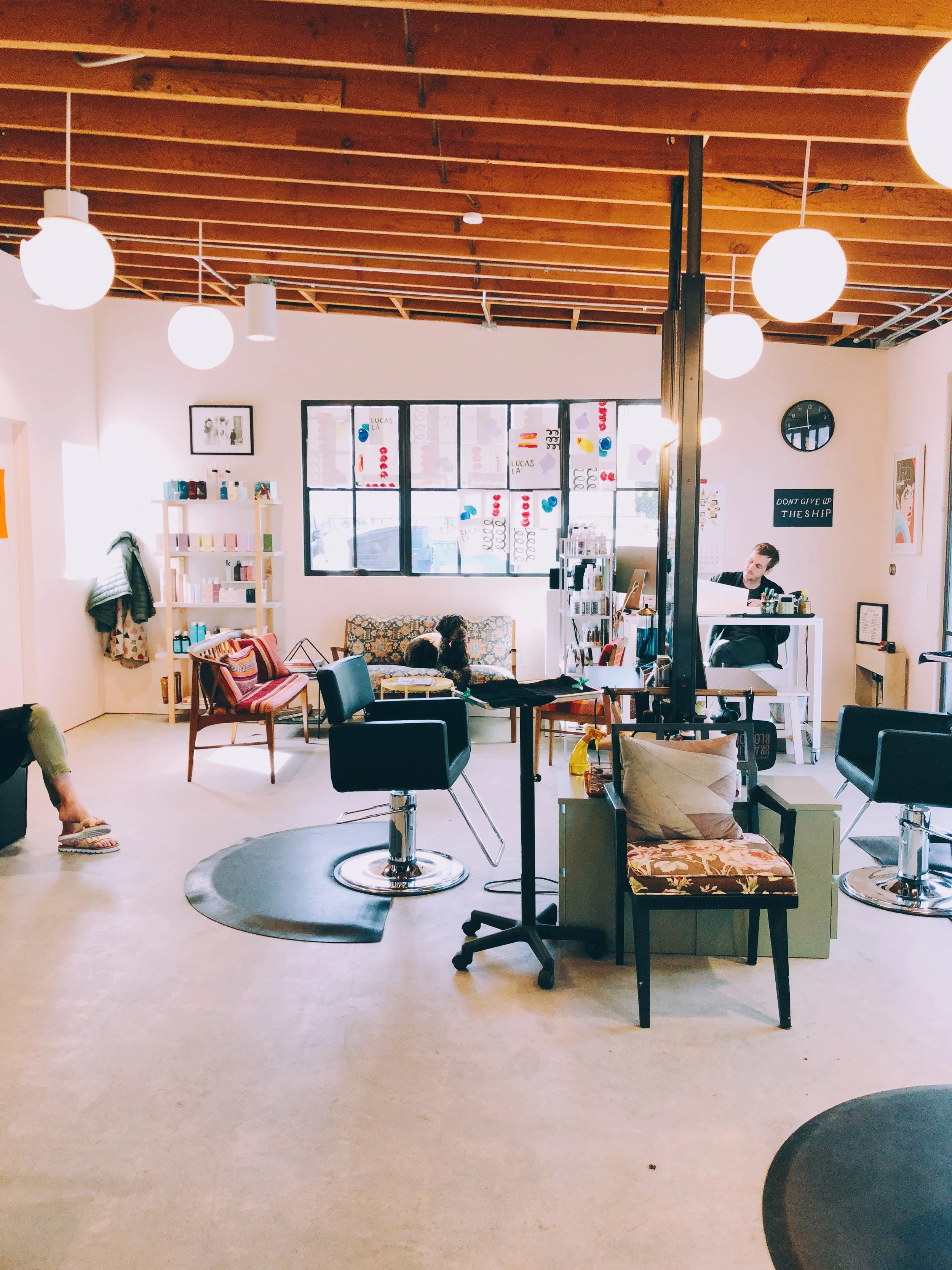 A cozy, well-lit interior space with modern decor, featuring chairs, sofas, shelves with products, artwork on the walls, and a person working at a desk.