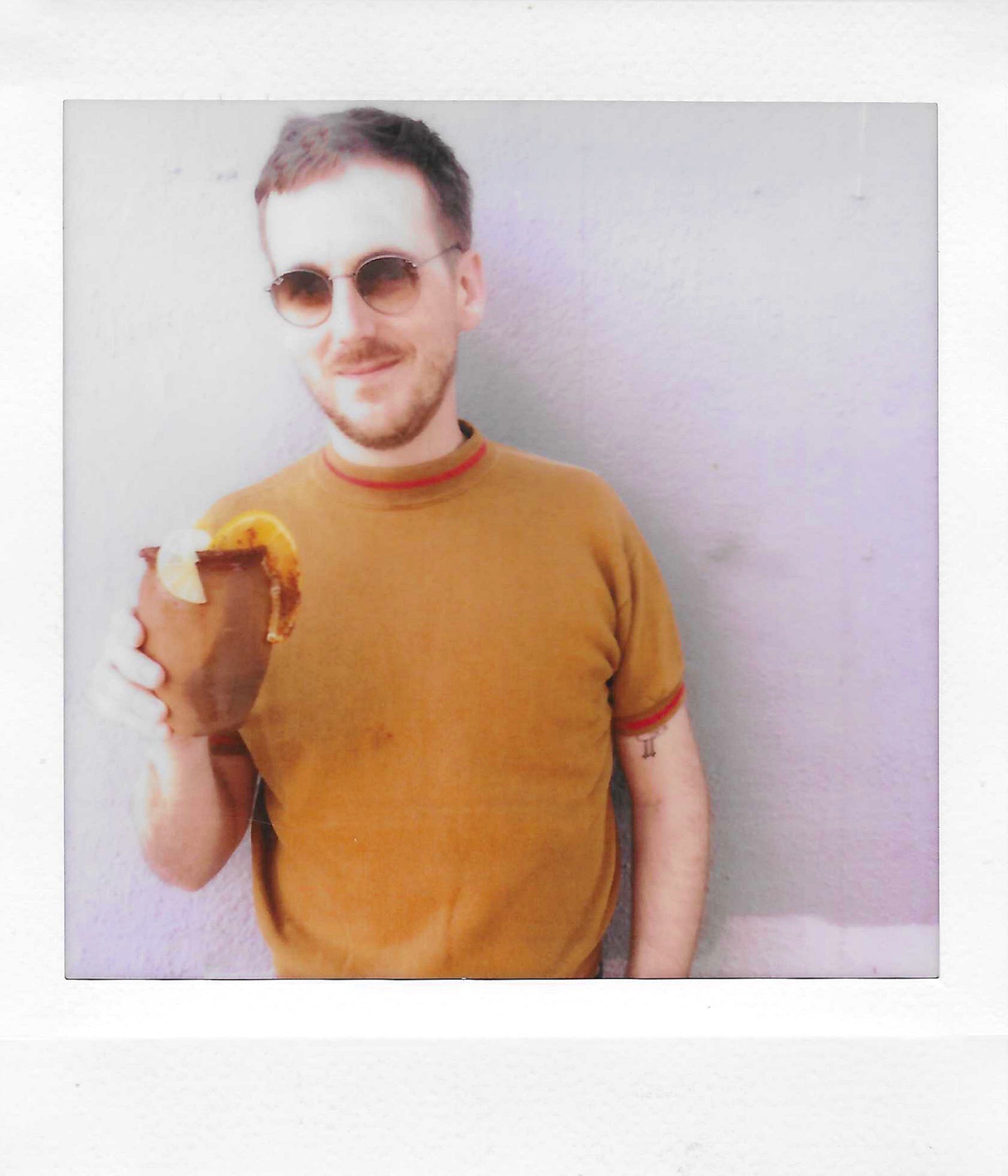 A young man with short dark hair, facial hair, and round sunglasses smiling and holding a slice of cake with icing and a fruit topping, wearing a mustard-colored shirt with red accents.