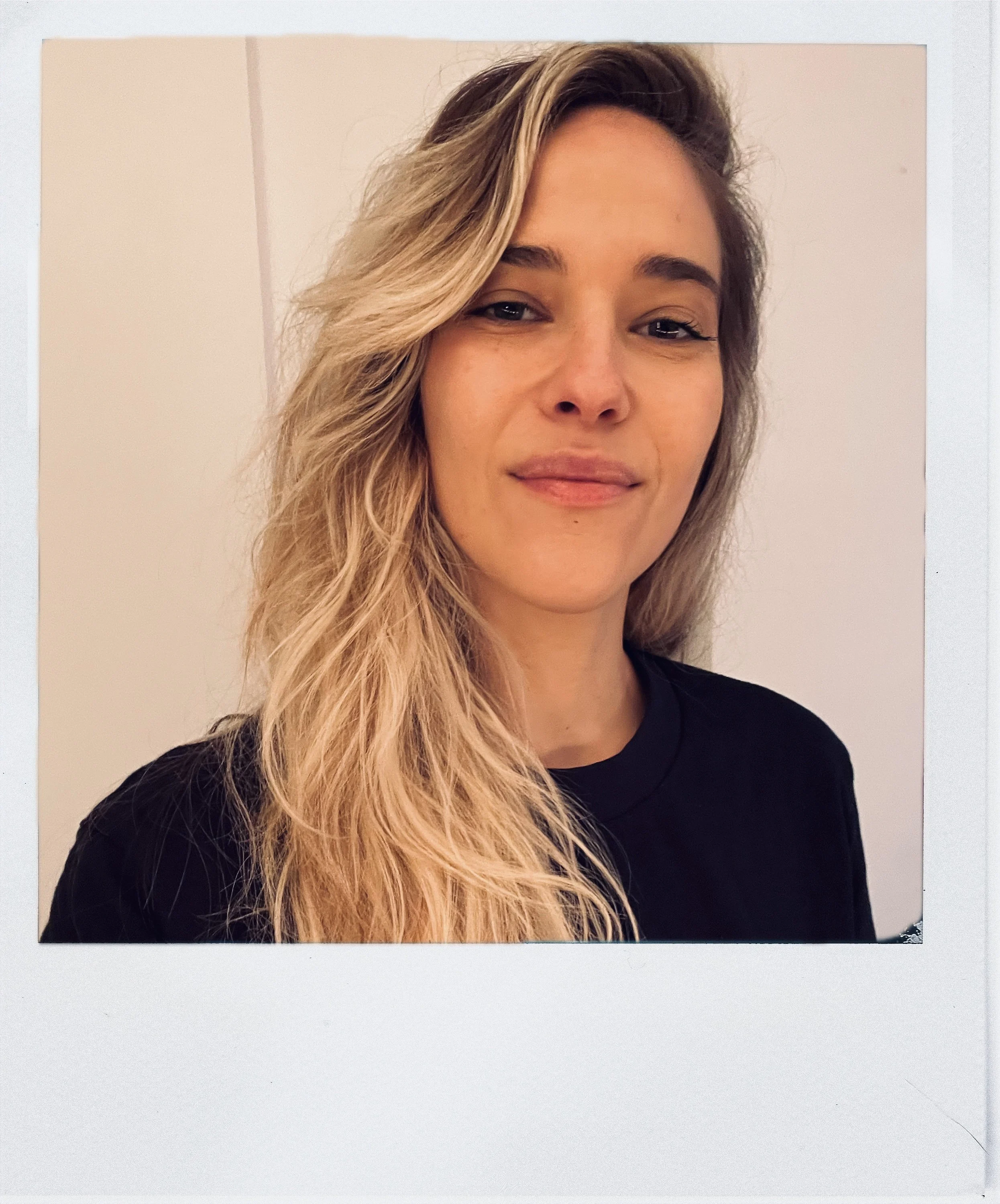 A woman with long wavy blonde hair is smiling slightly. She is wearing a black top and is against a pale wall background.