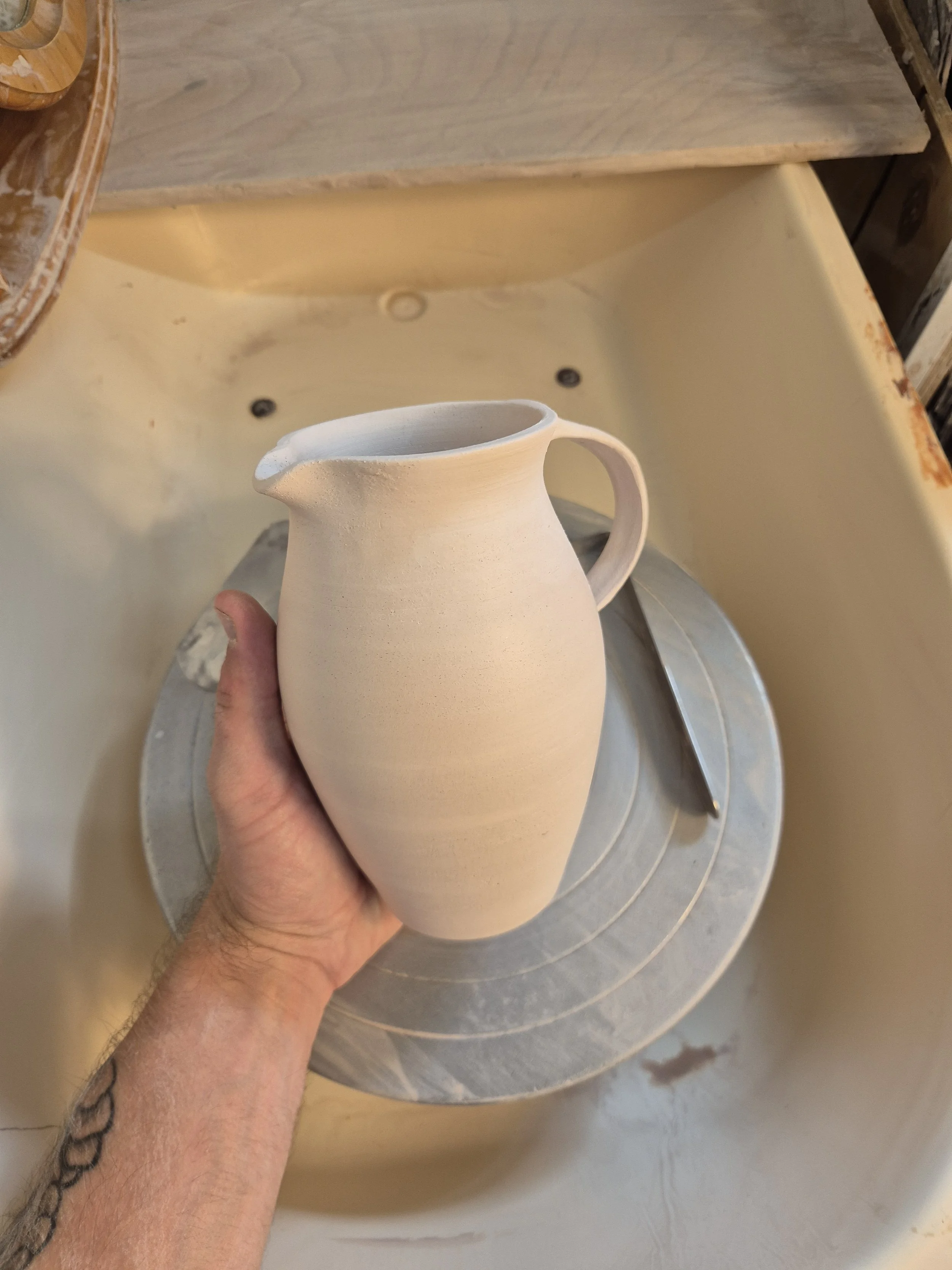 A person holding a white ceramic jug with a handle, in a pottery studio. The bowl-shaped wheel and a yellow wall are visible in the background.