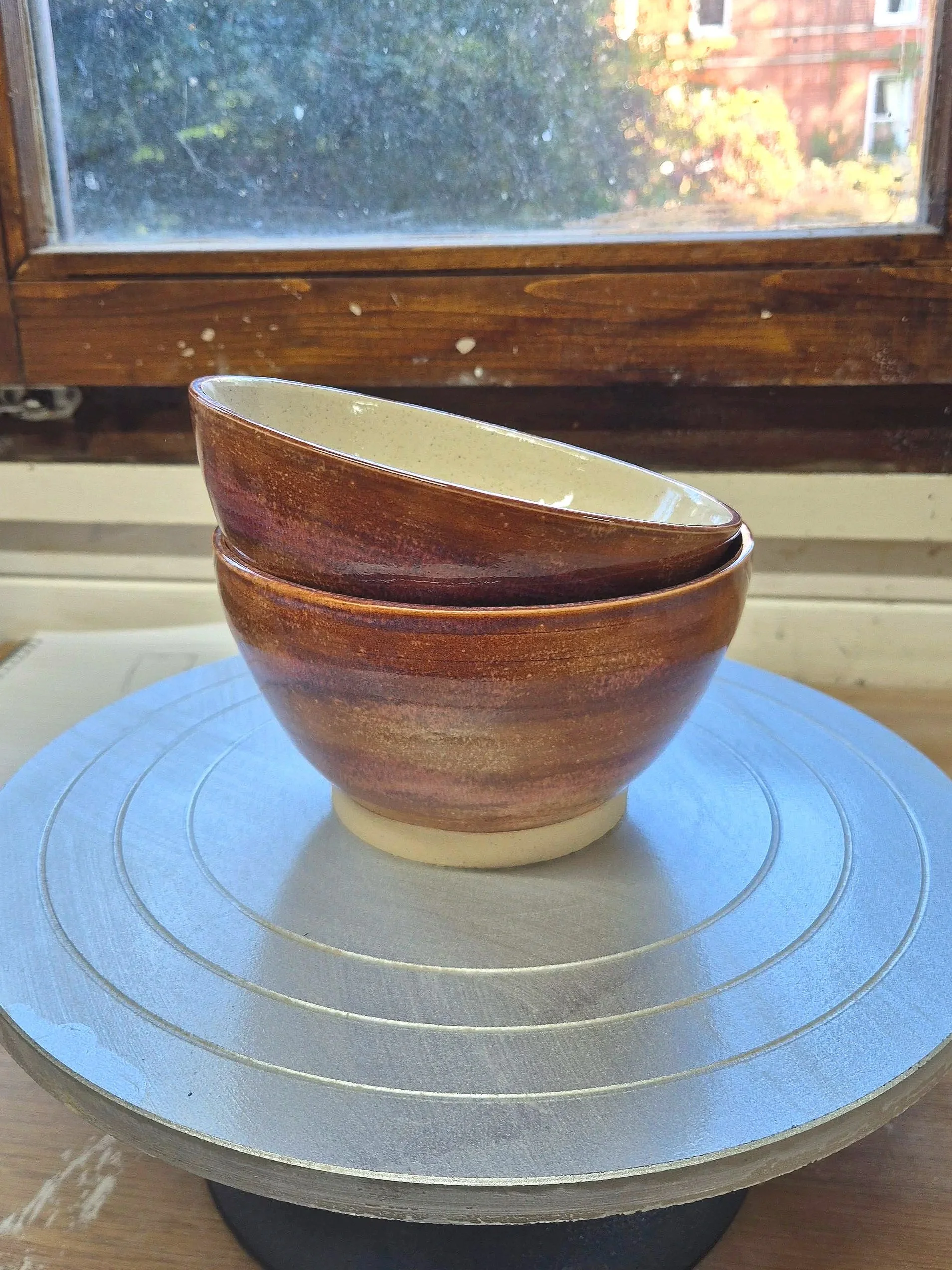 A ceramic bowl with a reddish-brown exterior and beige interior, stacked with another similar bowl, on a turntable in front of a window showing trees and a brick building outside.