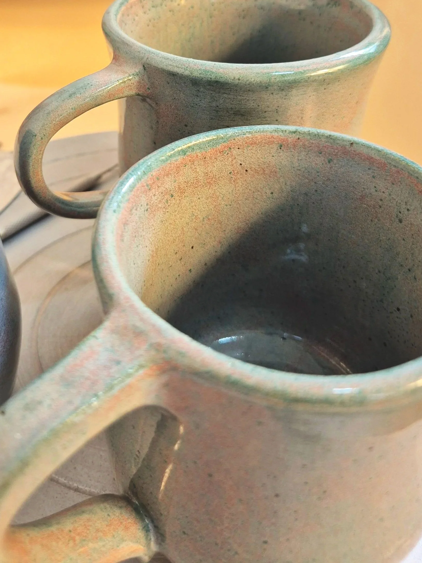 Close-up of two ceramic mugs with textured, multicolored glaze, placed on a surface with a yellow background.