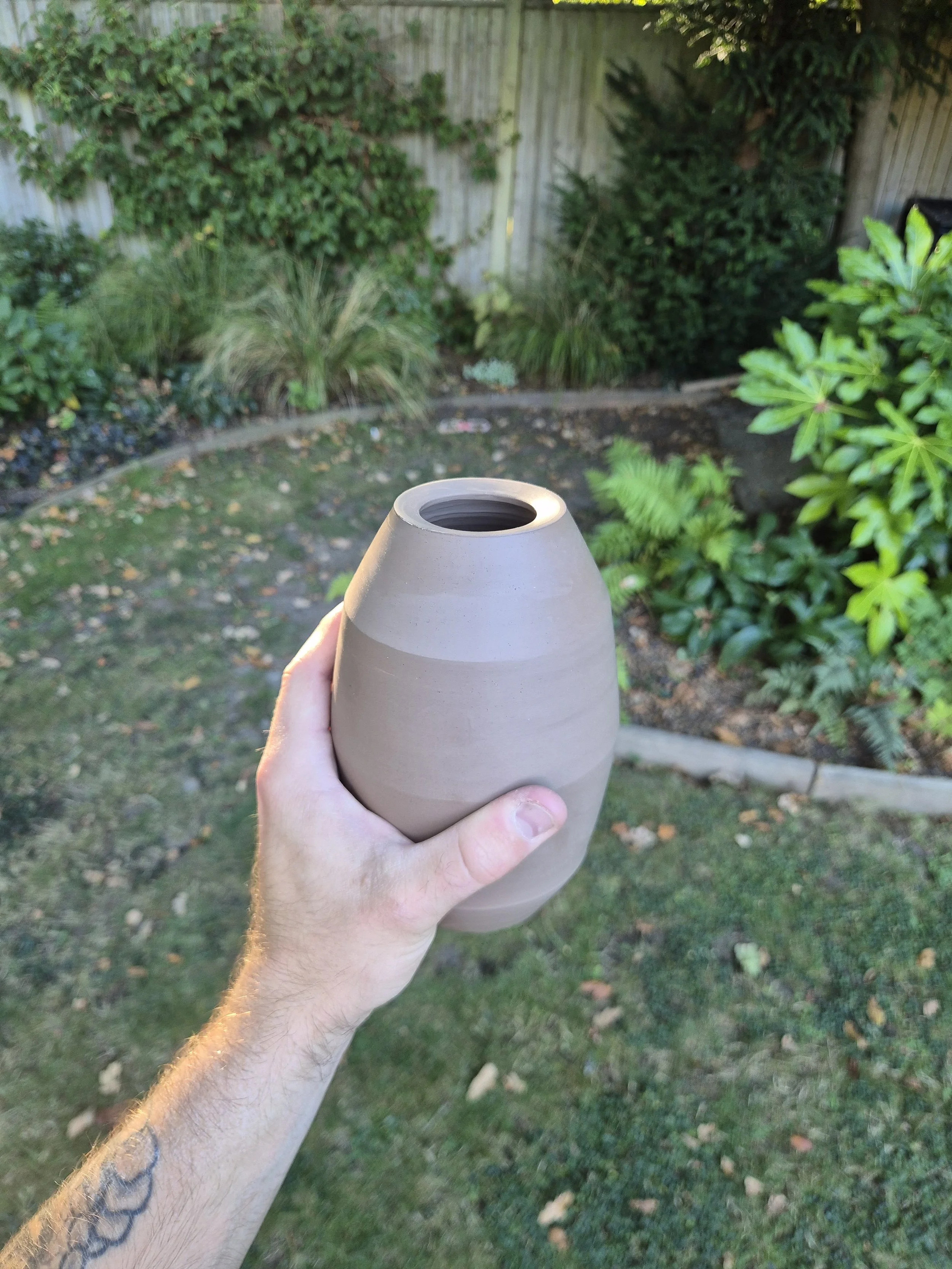 A hand holding a beige vase outdoors in a garden with green plants and a wooden fence in the background.