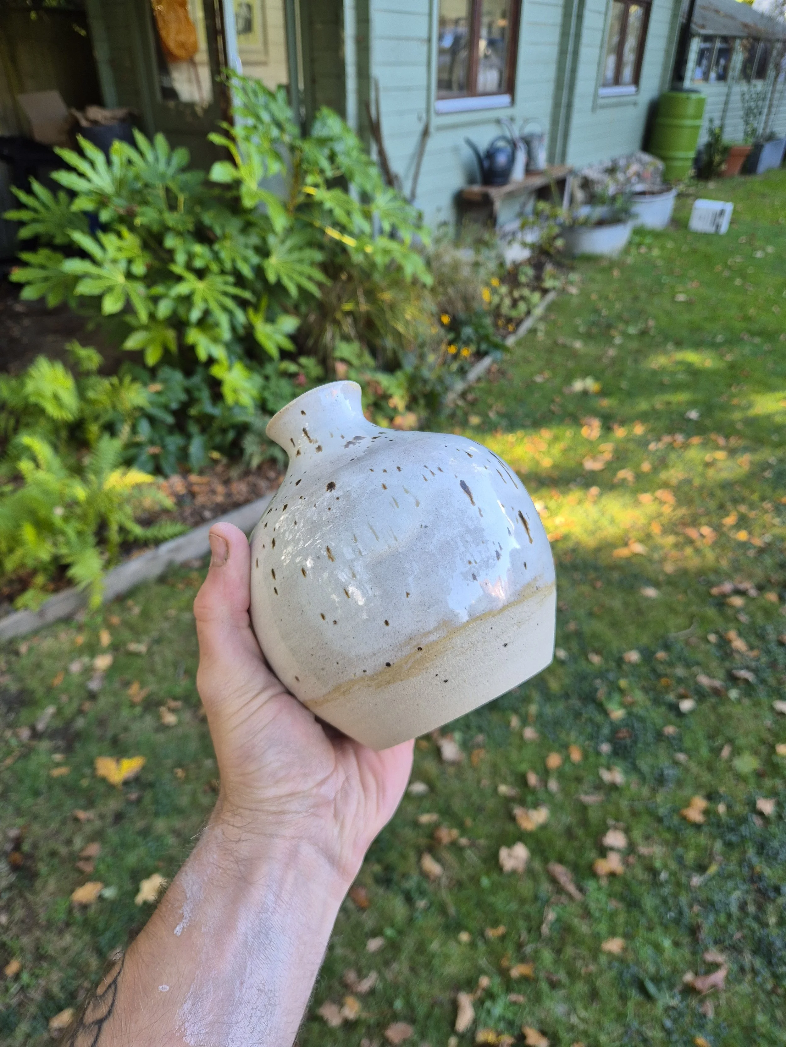 A person holding a beige ceramic vase outdoors in a garden with green plants and a house in the background.
