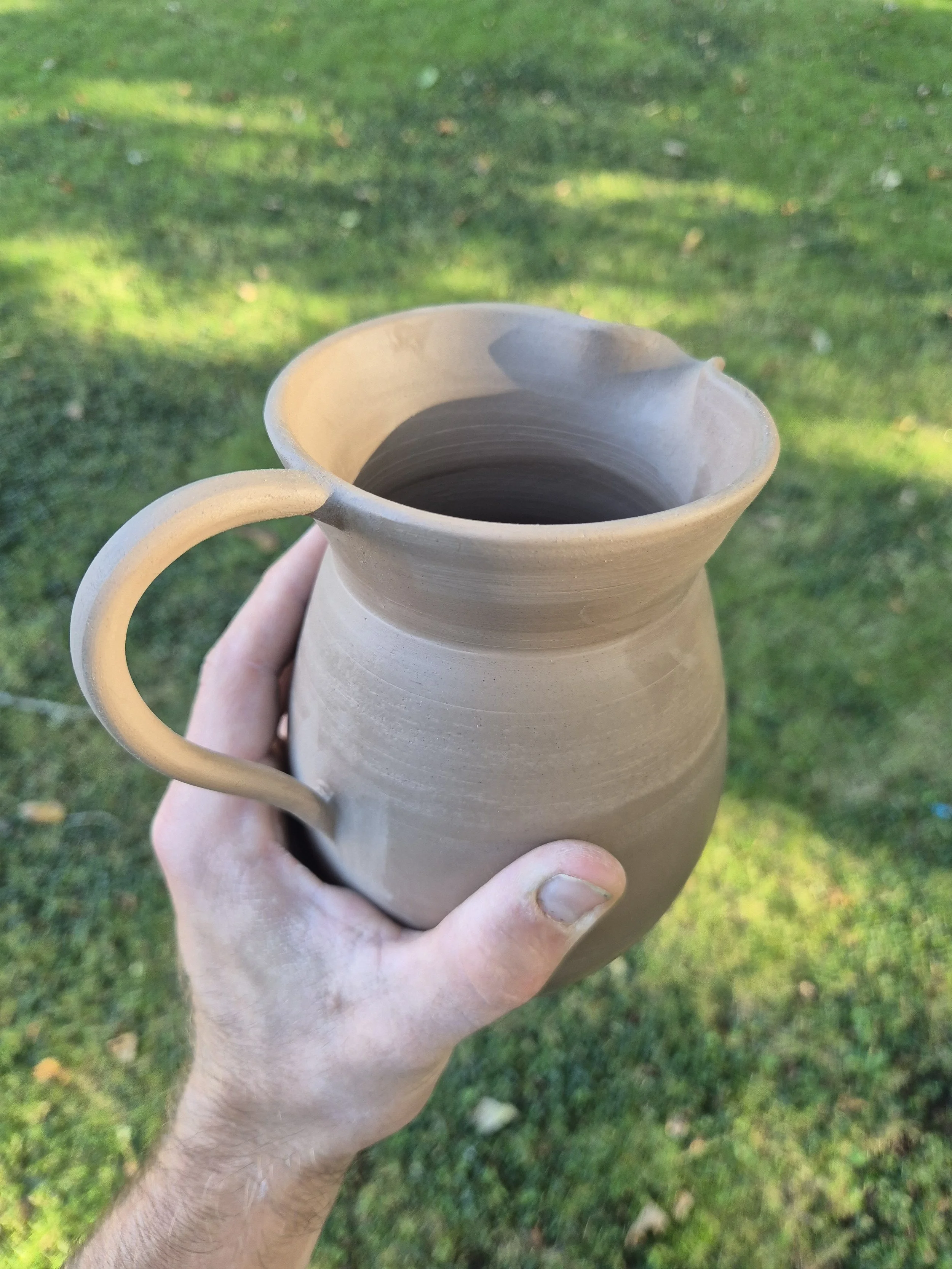 A person holding a beige ceramic pitcher with a handle, with a grassy outdoor background.