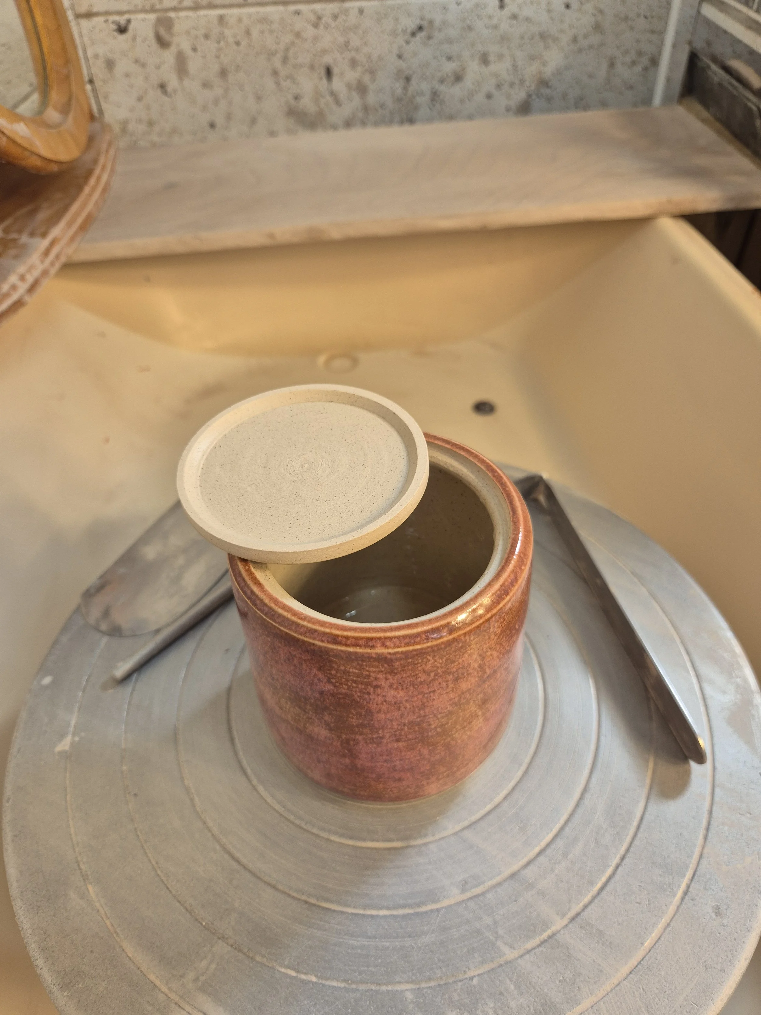 A pottery wheel with a partially finished ceramic mug on a turntable, a small sharpening stone, and a metal spatula nearby in a pottery studio.