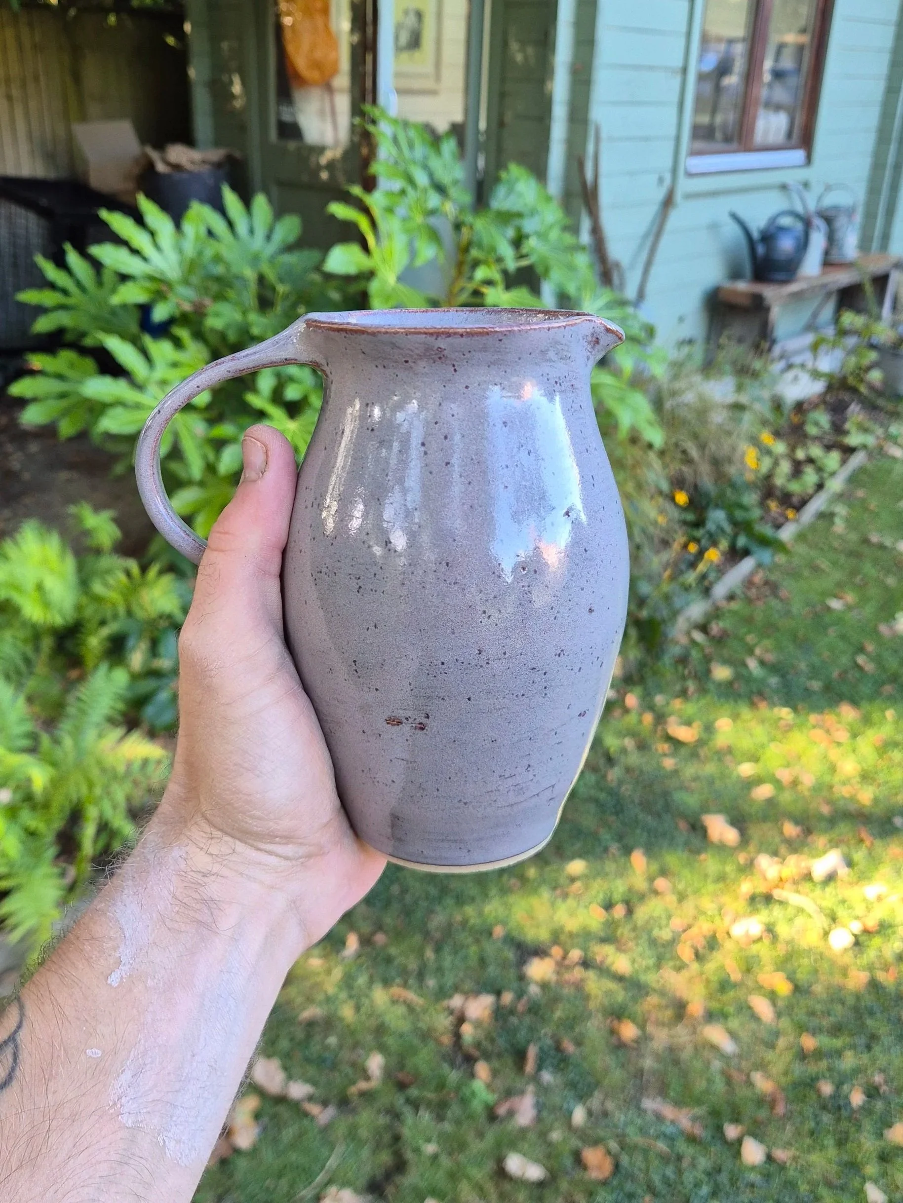 Person holding a decorated ceramic vase outdoors with greenery with a ceramic studio in the background