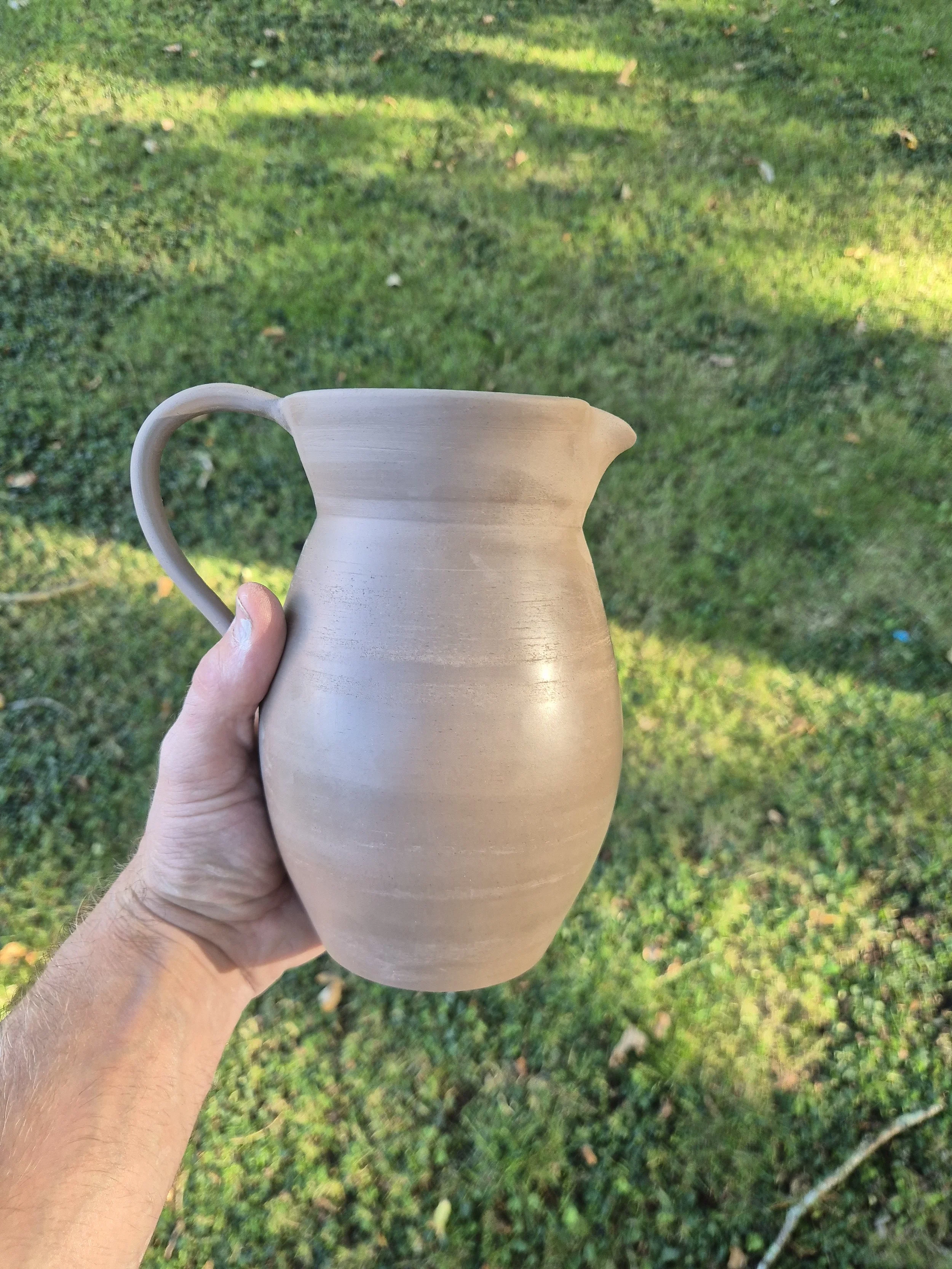 Hand holding a beige ceramic pitcher outdoors with grass in the background.