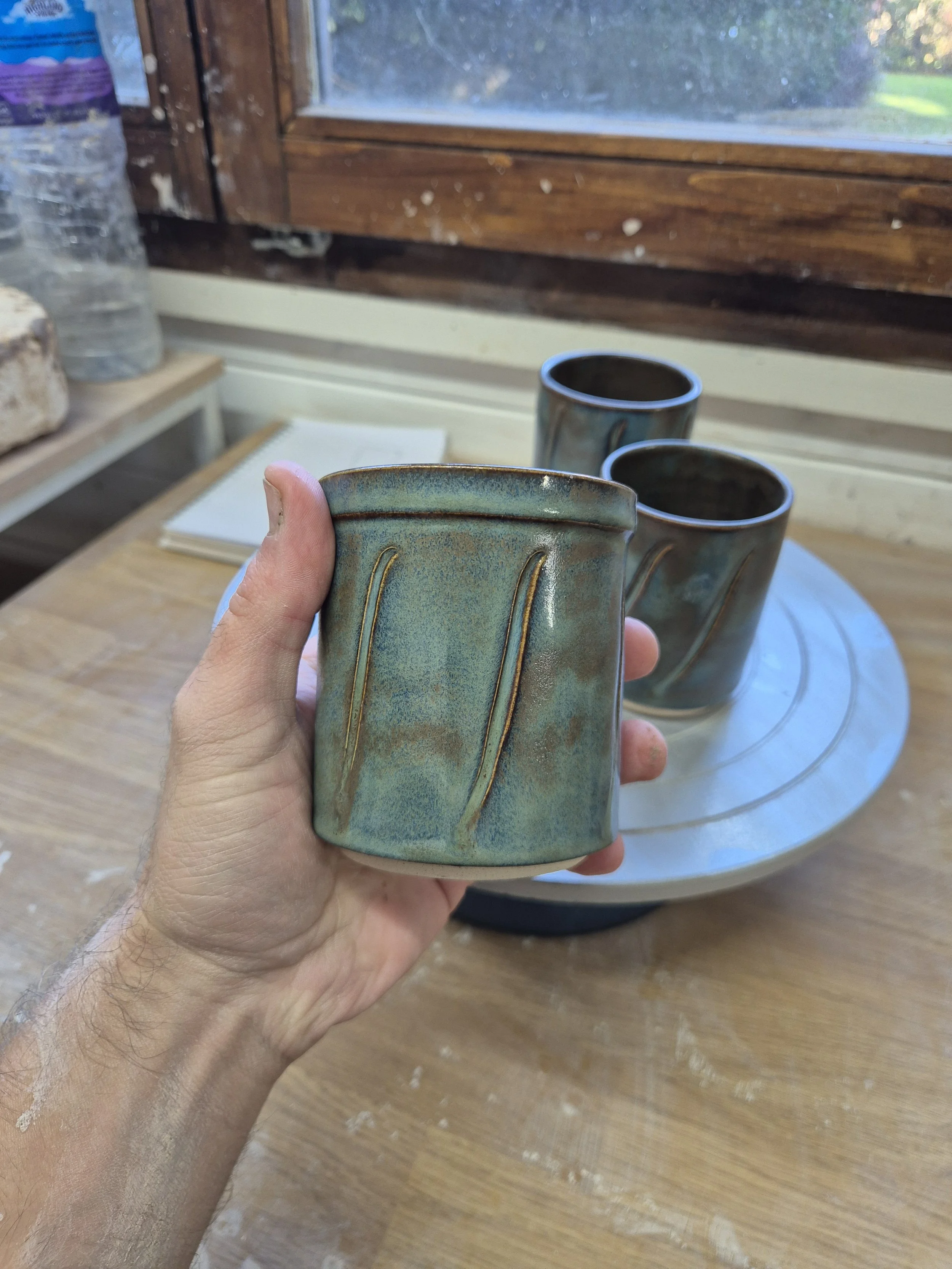 A hand holding three ceramic mugs in a room with a wooden table and a window in the background.