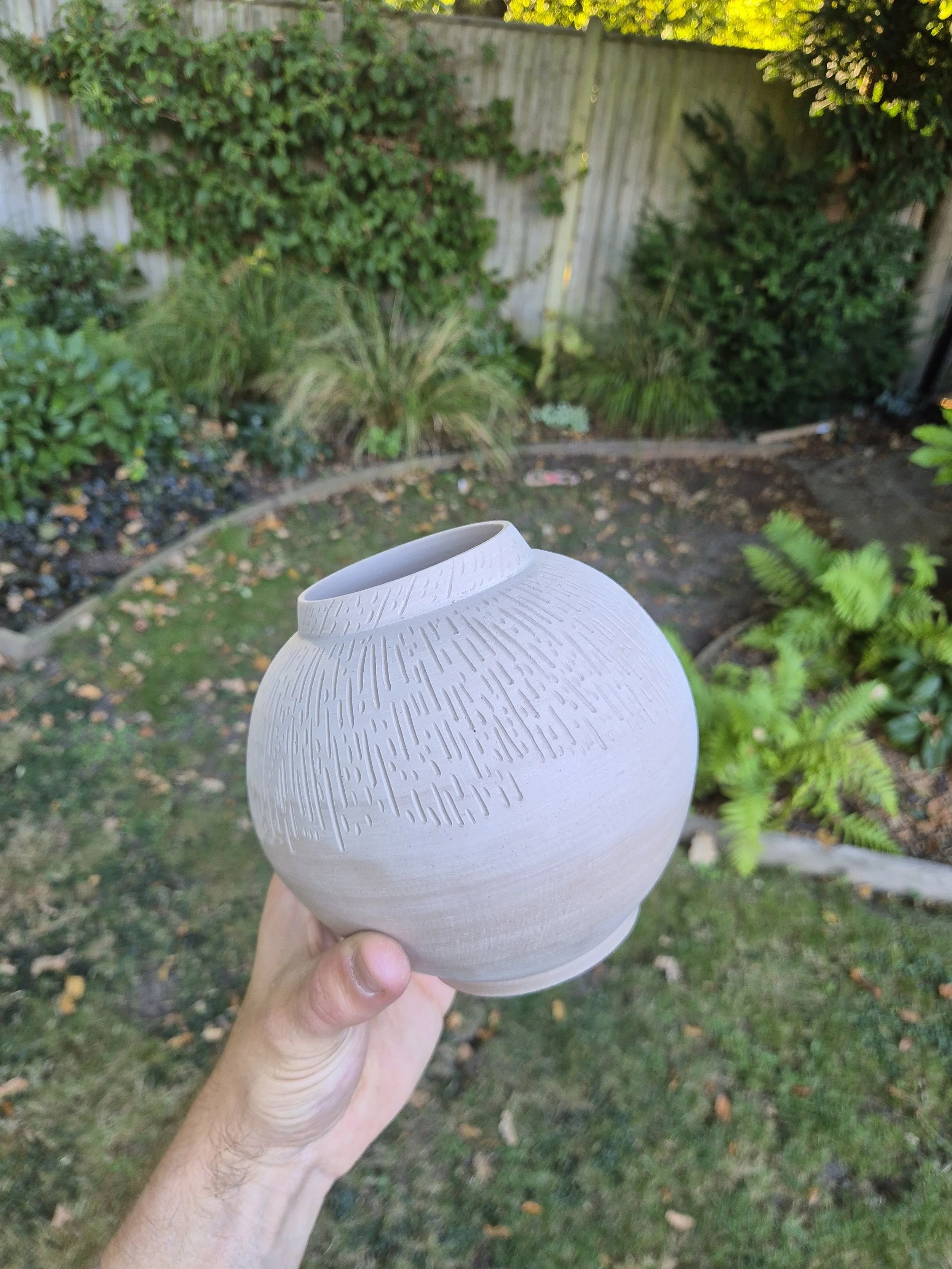 A person holding a white, textured, round ceramic vase outdoors in a garden with green bushes and trees in the background.