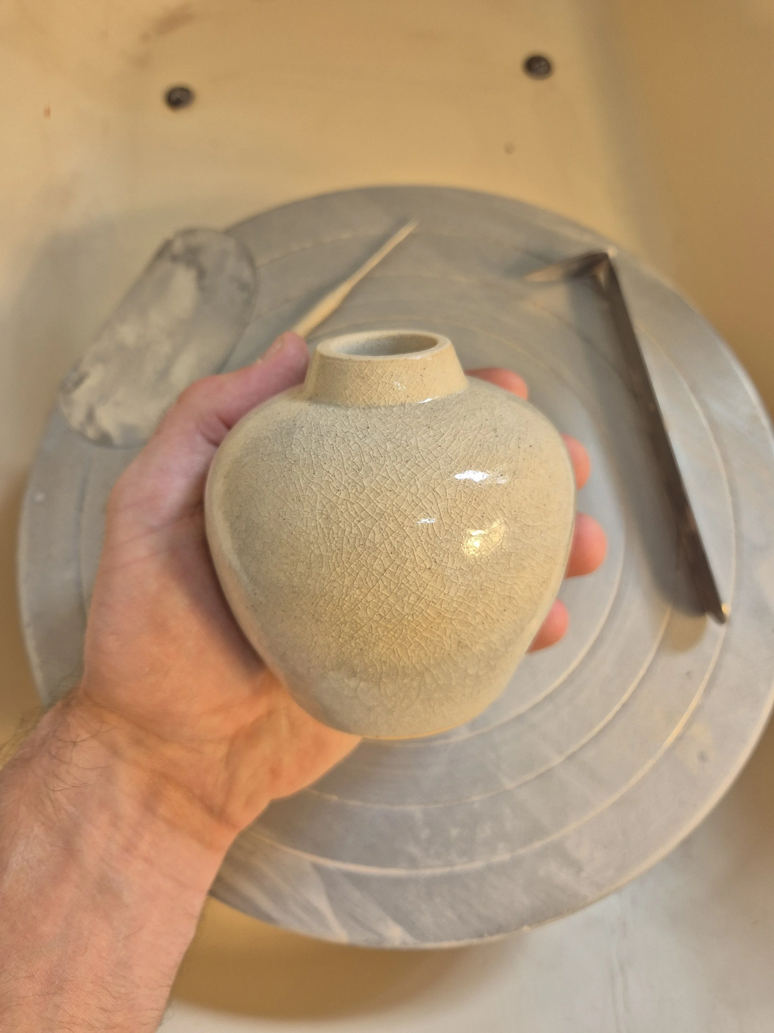 A person holding a small beige ceramic vase with a narrow opening in front of a pottery wheel in a studio.