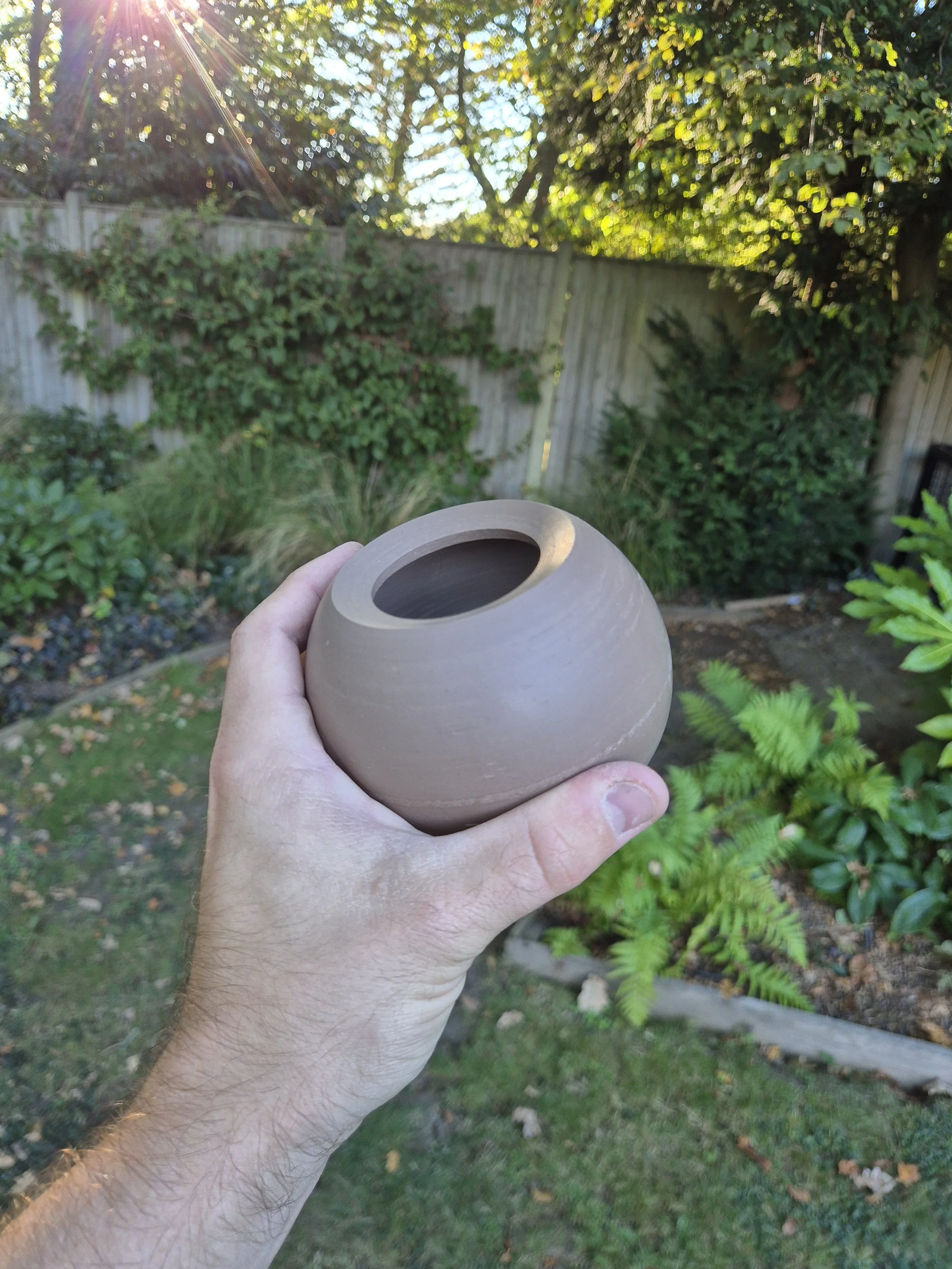 A person holding a round, light brown ceramic moon jar with a garden and a wooden fence in the background, sunlight shining through trees.