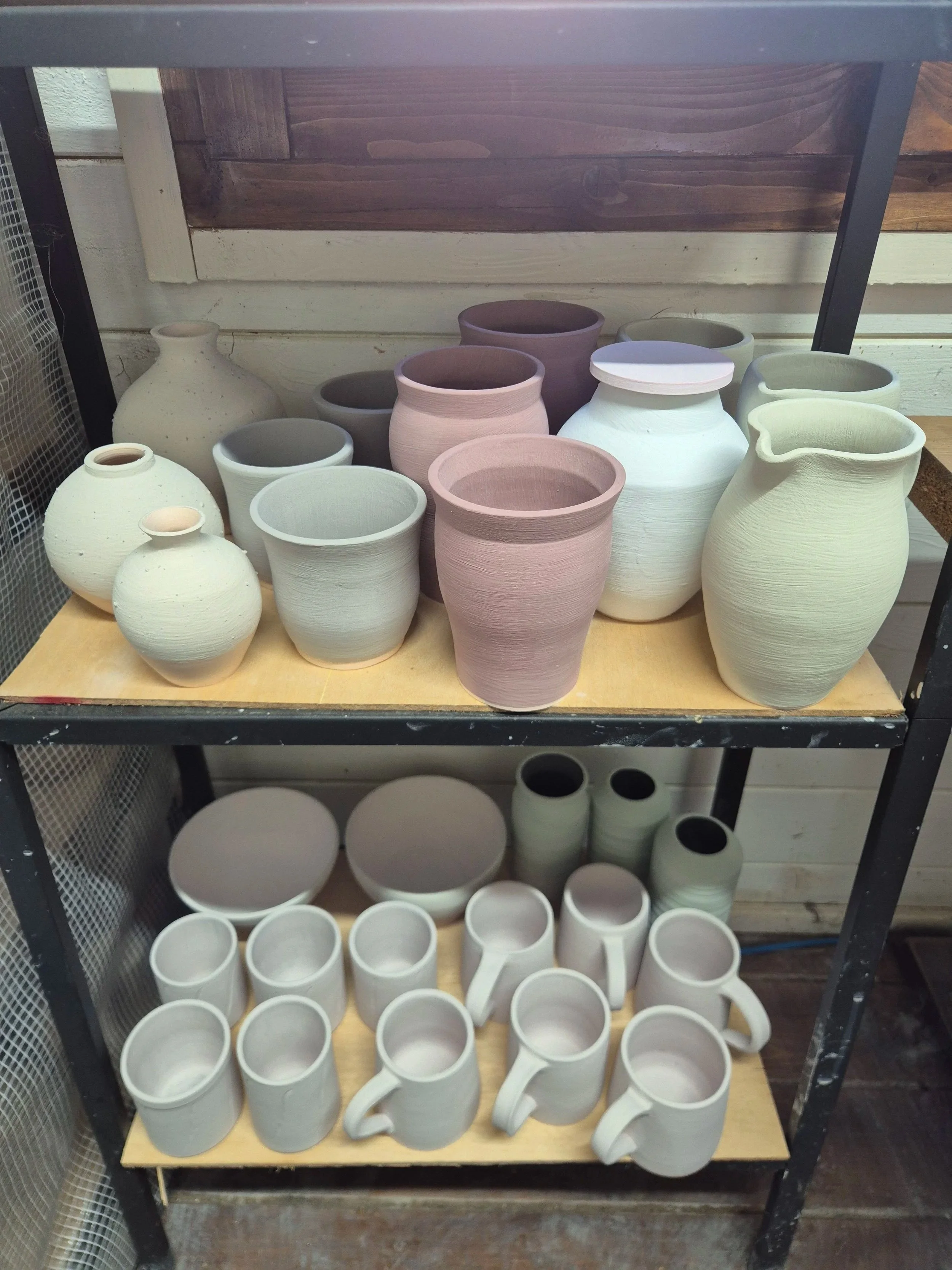 Shelving unit with a variety of ceramic flowerpots and mugs, most of which are white or light-colored, arranged on two shelves, with wooden and metal framing in the background. all pots ready to be glazes