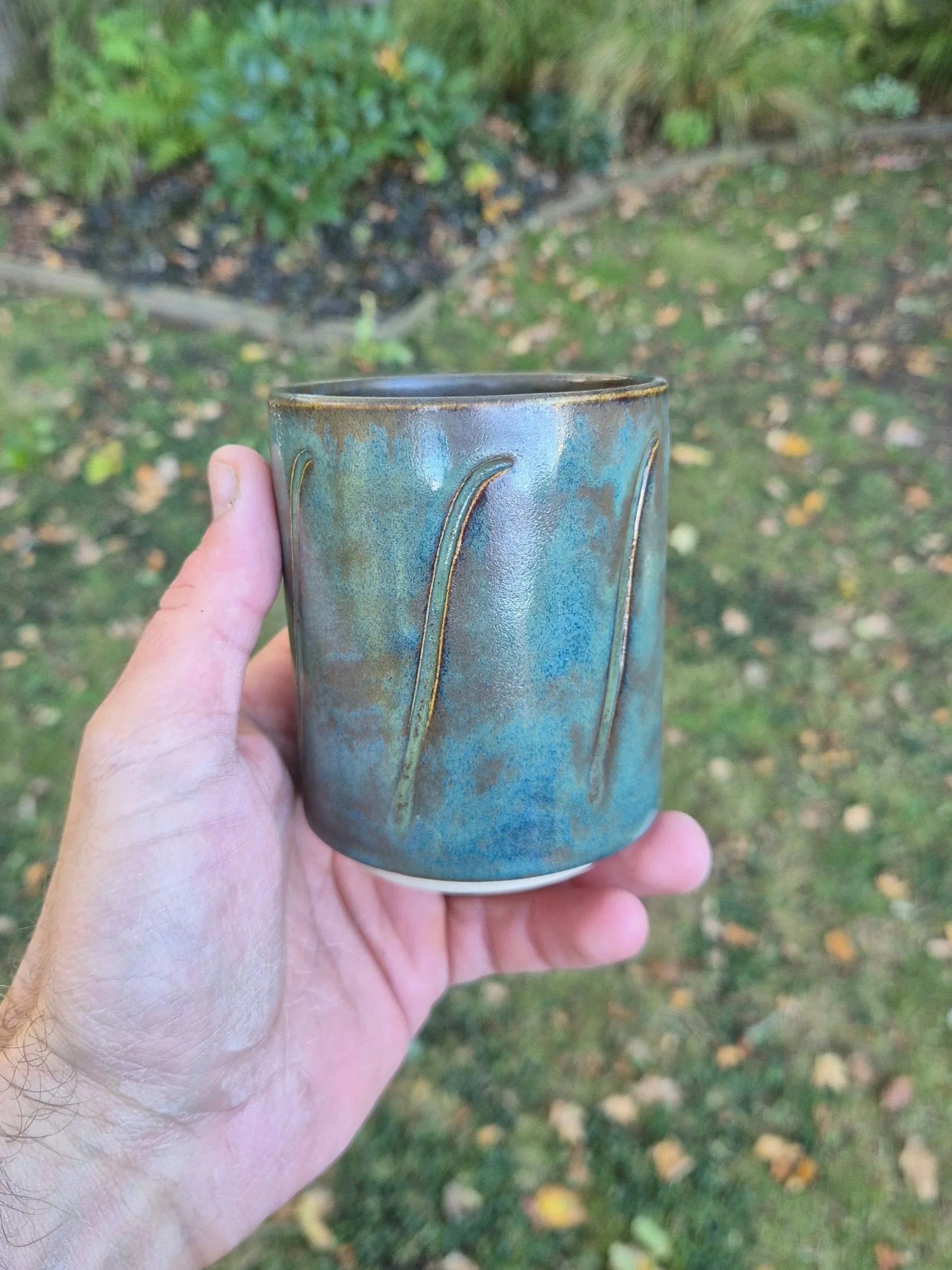 Person holding a handmade ceramic mug outdoors with greenery and fallen leaves in the background.