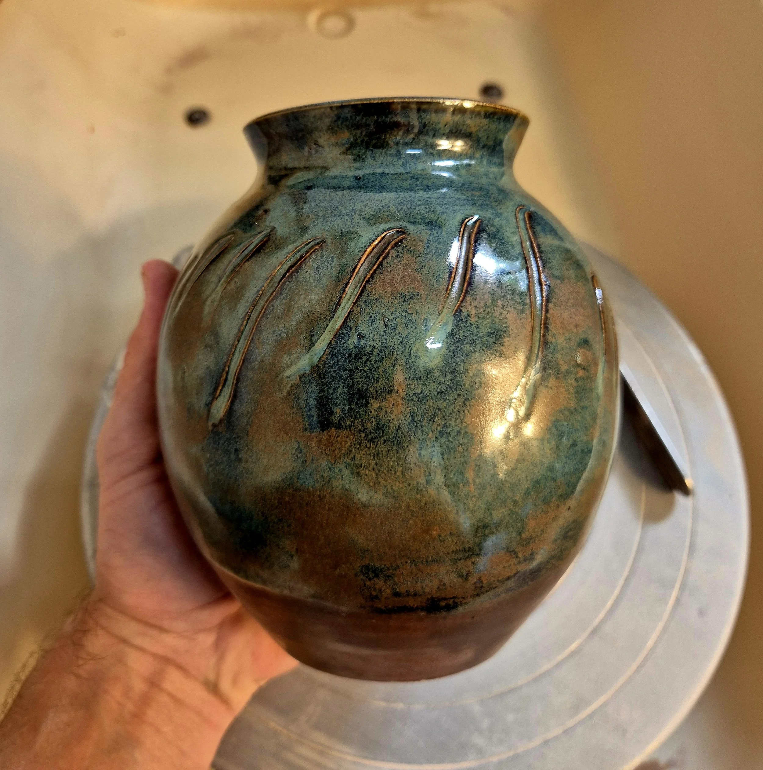 Close-up of a ceramic vase with a textured green and brown glaze, held by a person's hand.