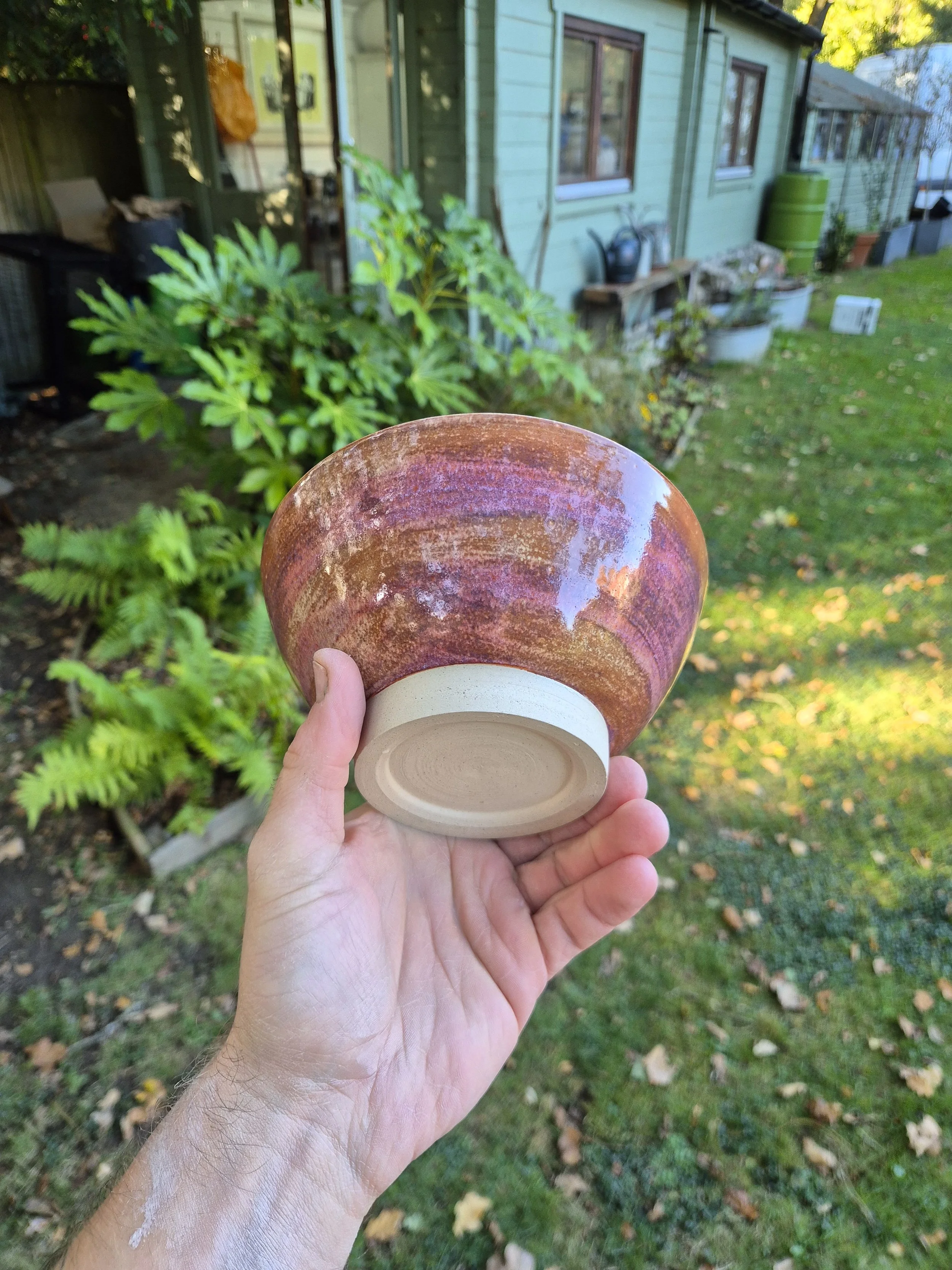 A person holding a ceramic bowl outdoors in front of a green house with plants and garden items in the background.