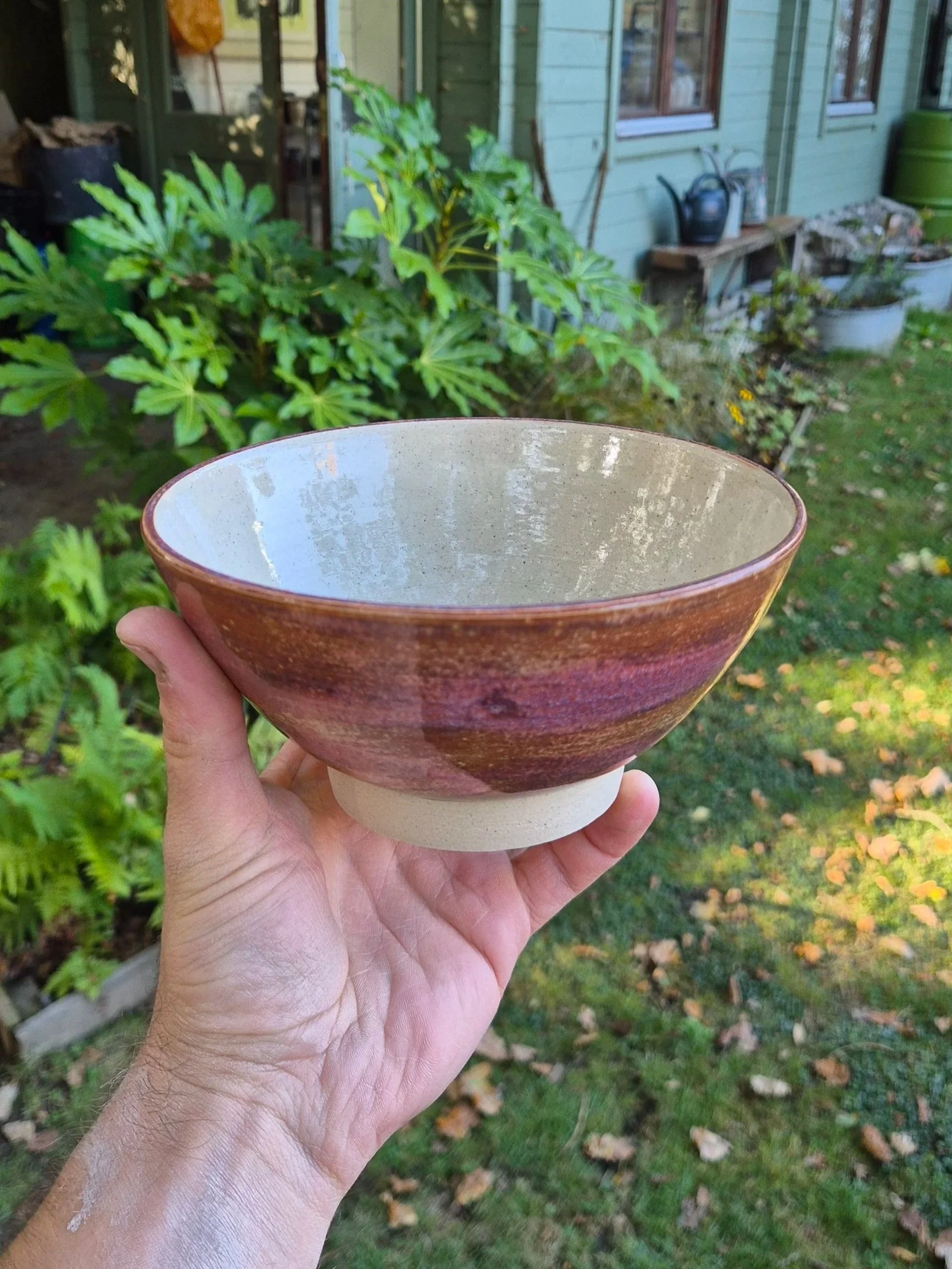 A person holding a ceramic bowl outdoors with a green garden, plants, with a ceramic studio in the background