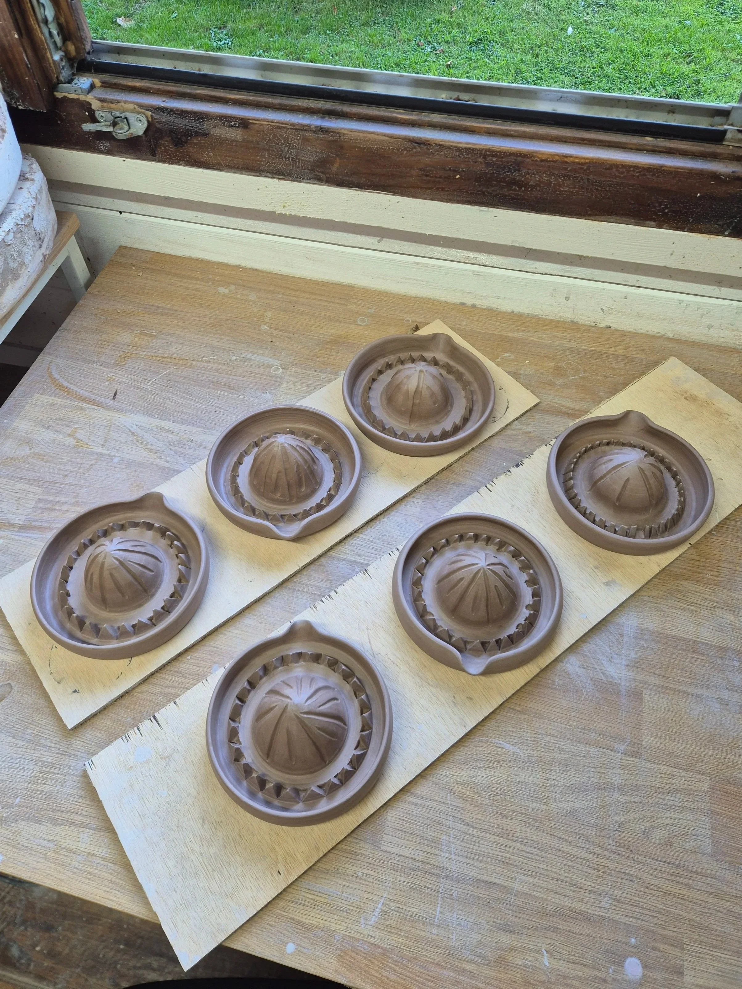 Unfinished ceramic citrus juicer molds on a wooden table near a window, with a grassy lawn outside.
