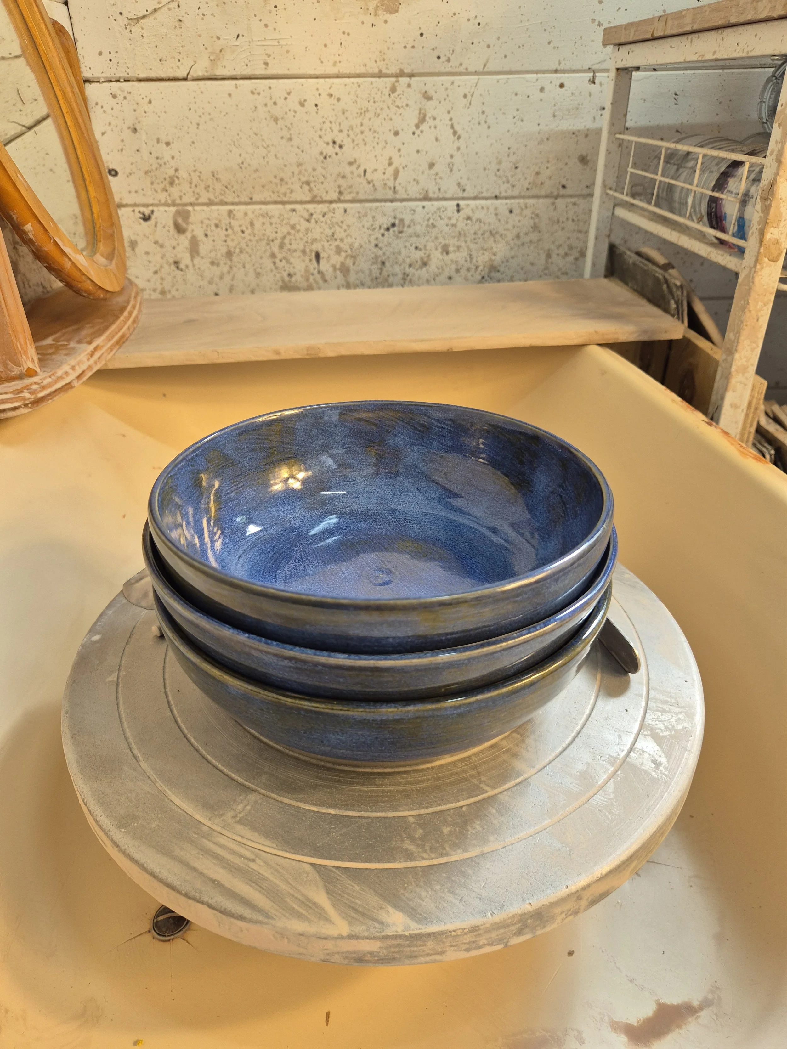 A blue ceramic bowl on a spinning wheel in a pottery studio, with shelves and wooden storage in the background.