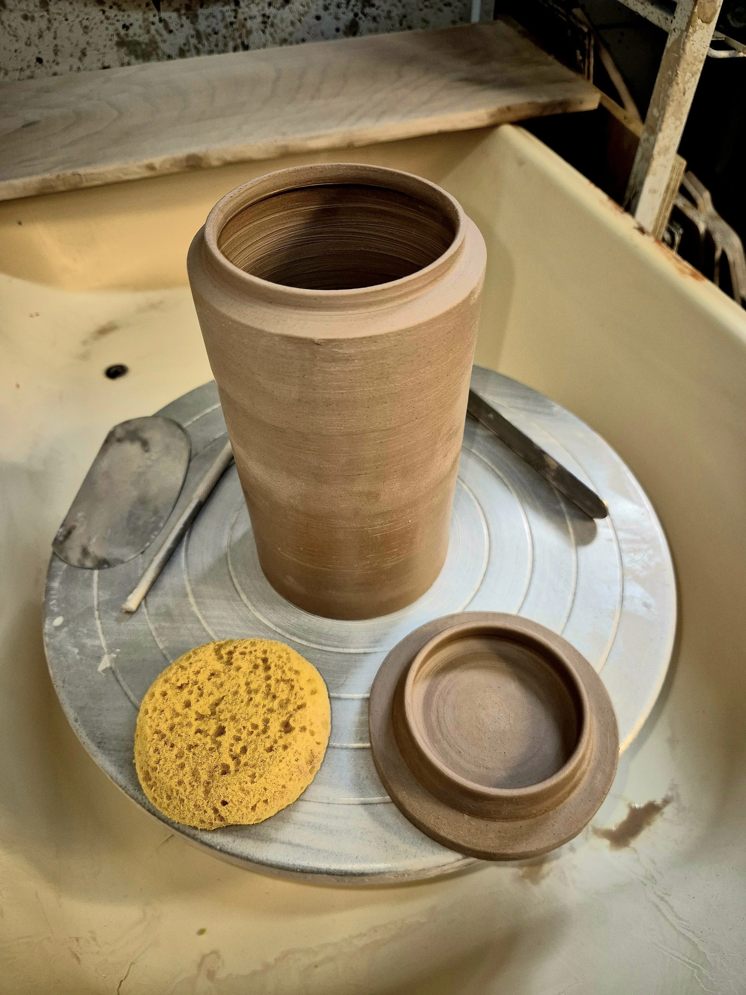 A pottery wheel with a partially formed clay vase, a sponge, and a small bowl on it, in a pottery studio.