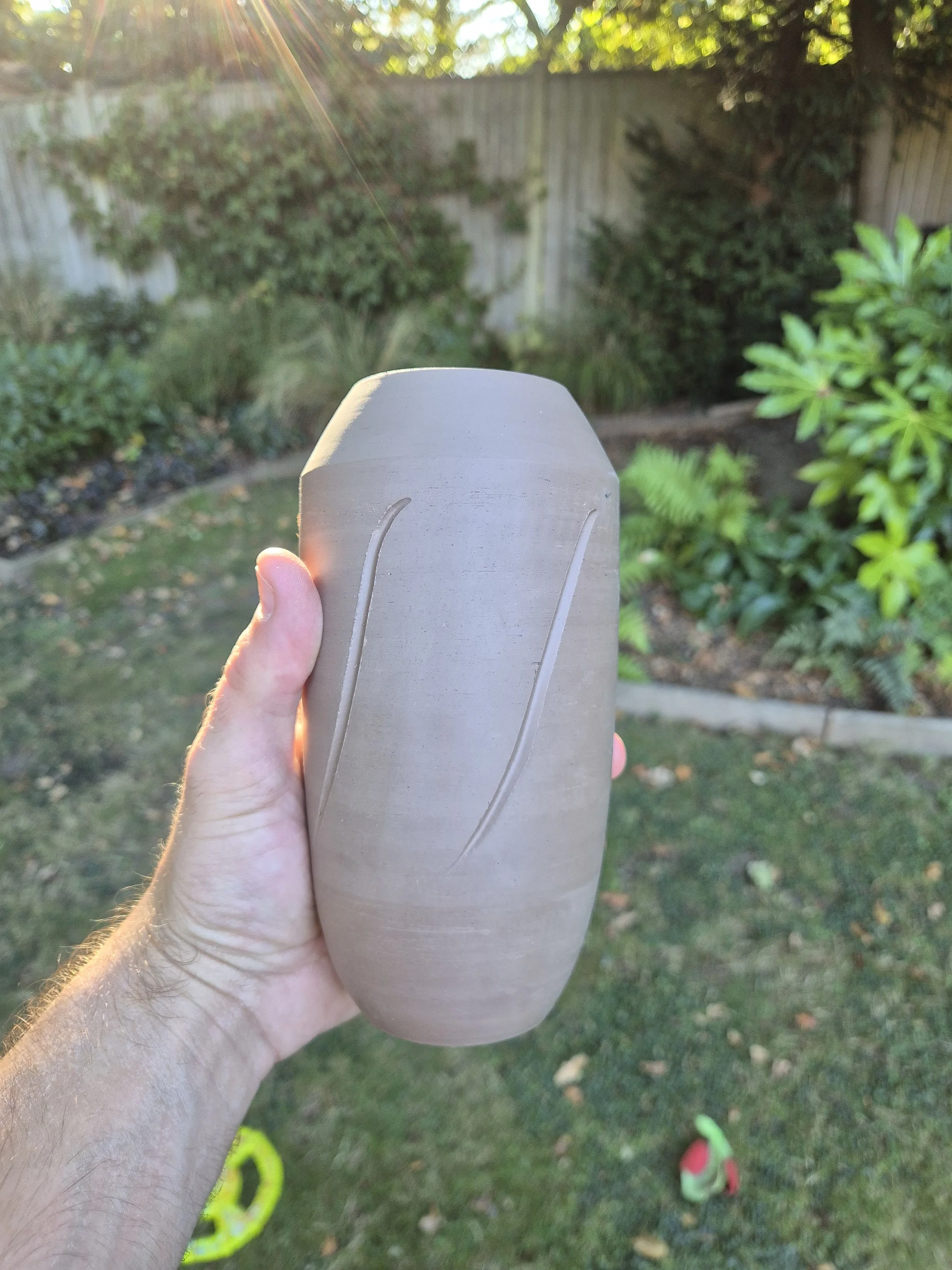 Person holding a vase outdoors in a backyard with grass, bushes, and a wooden fence in the background, with sunlight shining.