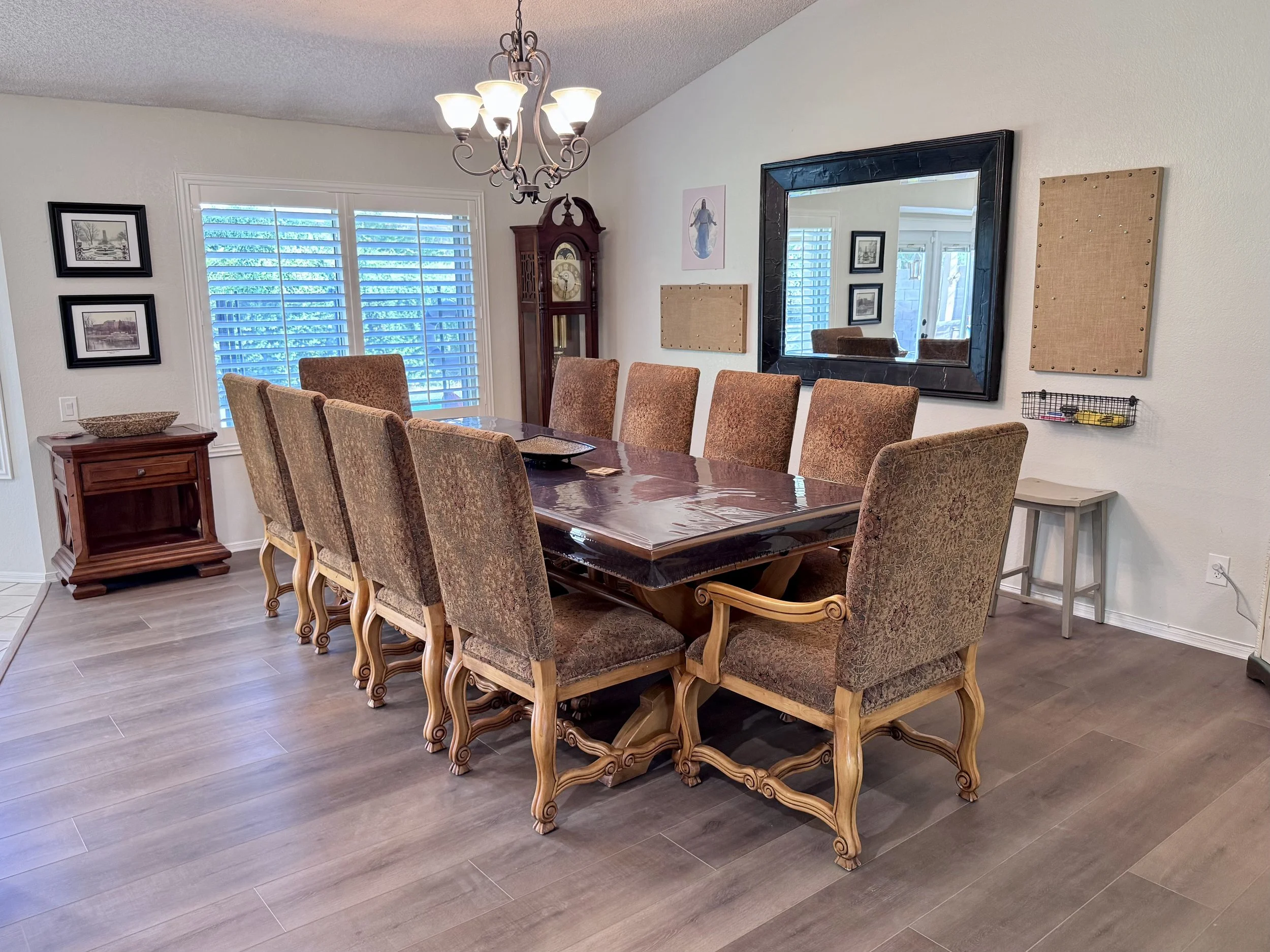 A dining room with a long dark wood table surrounded by ornate upholstered chairs. There is a wooden side table with a wicker basket and a tall grandfather clock in the corner. The wall features a large black-framed mirror, small framed pictures, and bulletin boards. A window with white shutters and a chandelier hangs from the ceiling, illuminating the room.