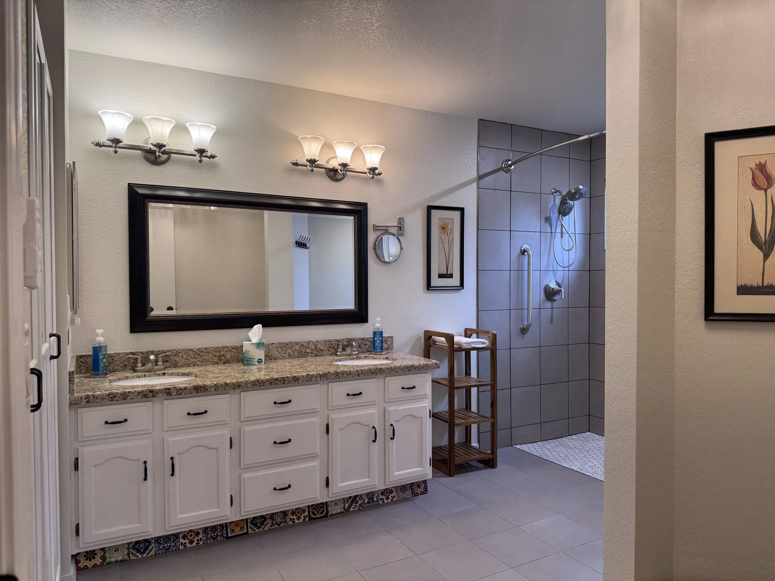A bathroom with a double sink vanity, large mirror, framed art, and a shower area with gray tiles, a sliding shower curtain rod, and a small wooden shelving unit.