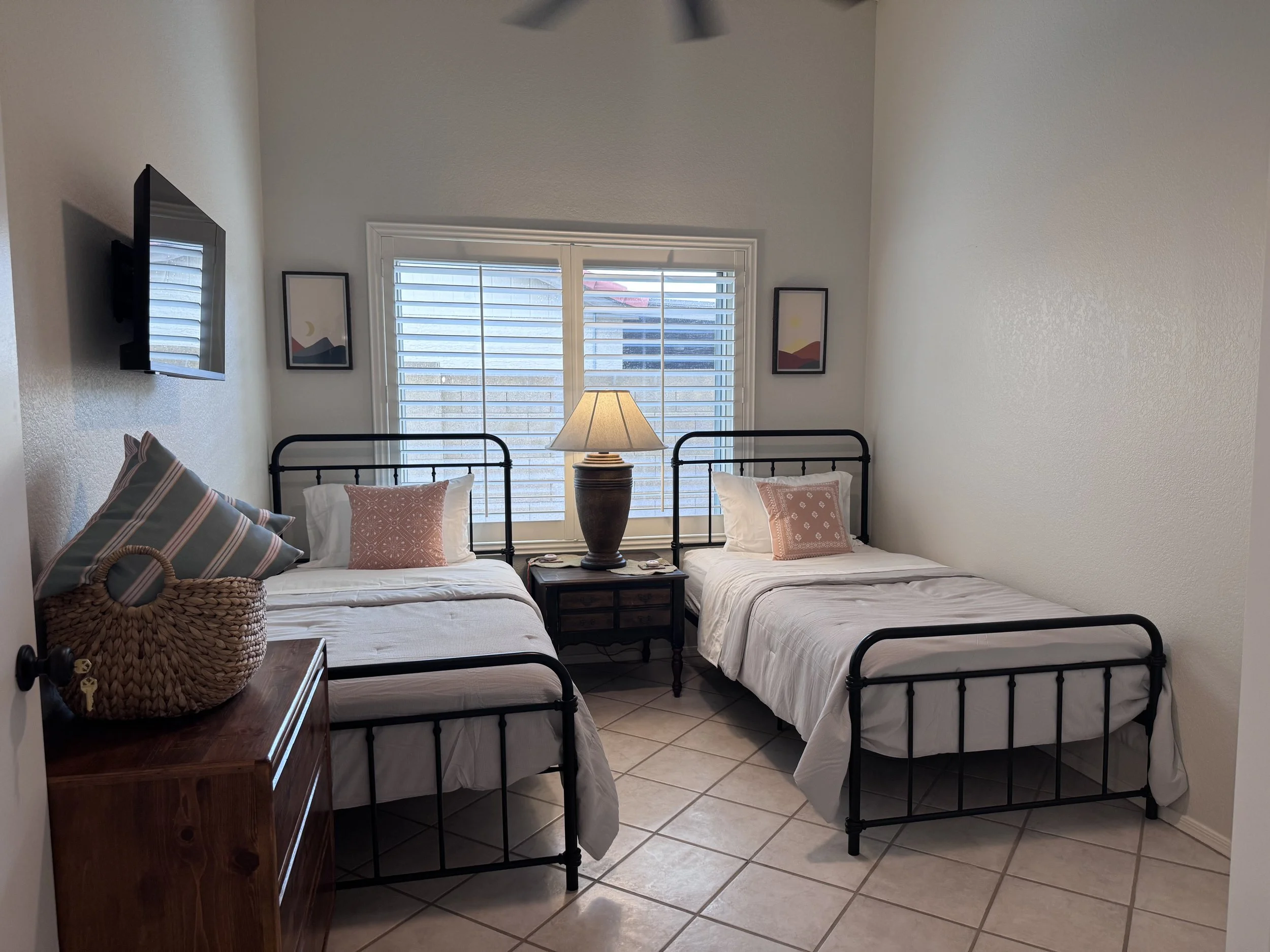 Two beds with black metal frames in a bedroom, each with white bedding and decorative pillows. A wooden nightstand with a lamp sits between the beds, with a window with white blinds behind it. There are two framed landscape pictures on the wall beside the window, and a flat-screen TV is mounted on the wall to the left.