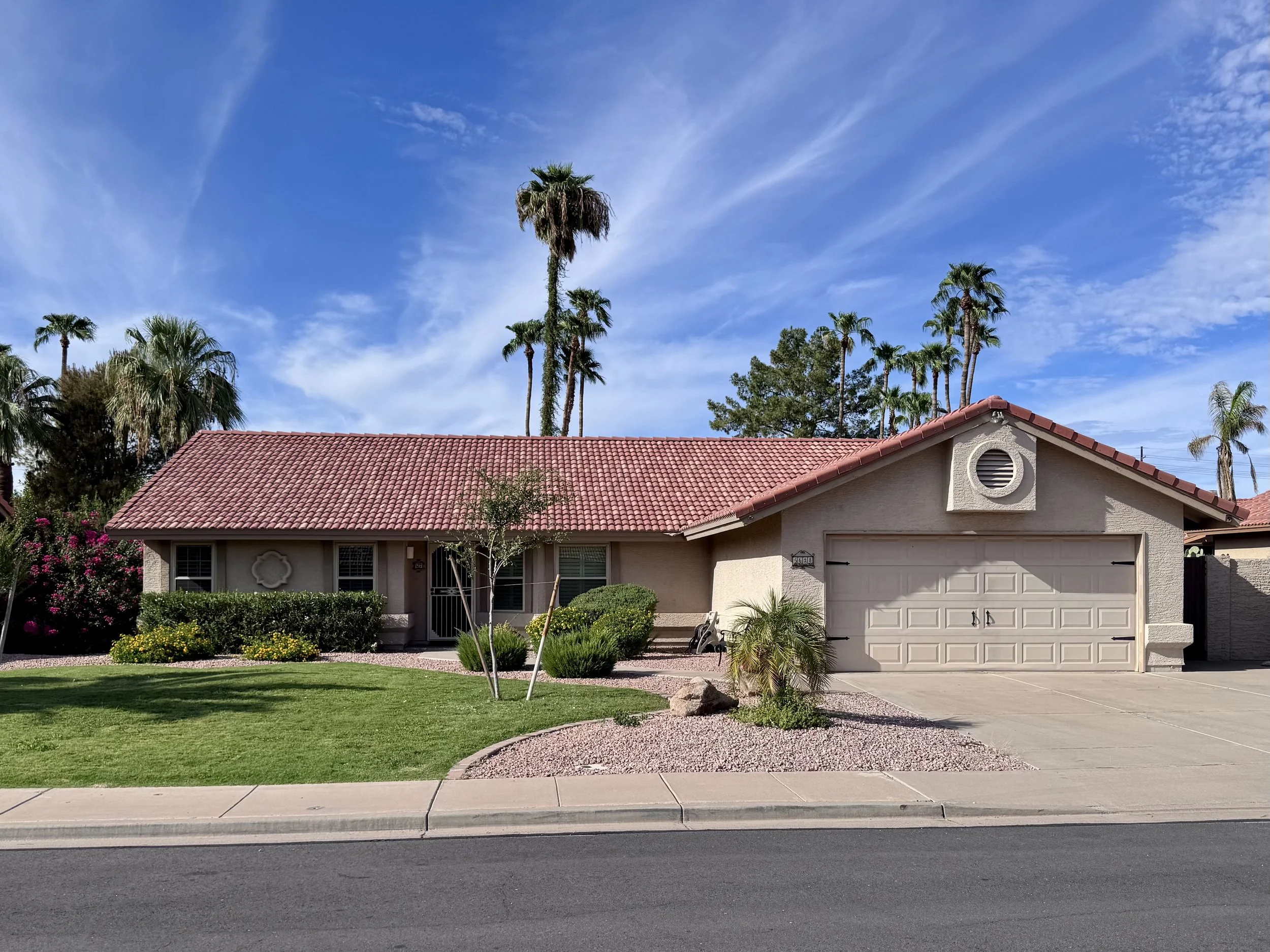 A single-story house with a red tile roof and a beige exterior is surrounded by a green lawn, bushes, and small trees. Tall palm trees are visible behind the house, and a blue sky with some wispy clouds is overhead.