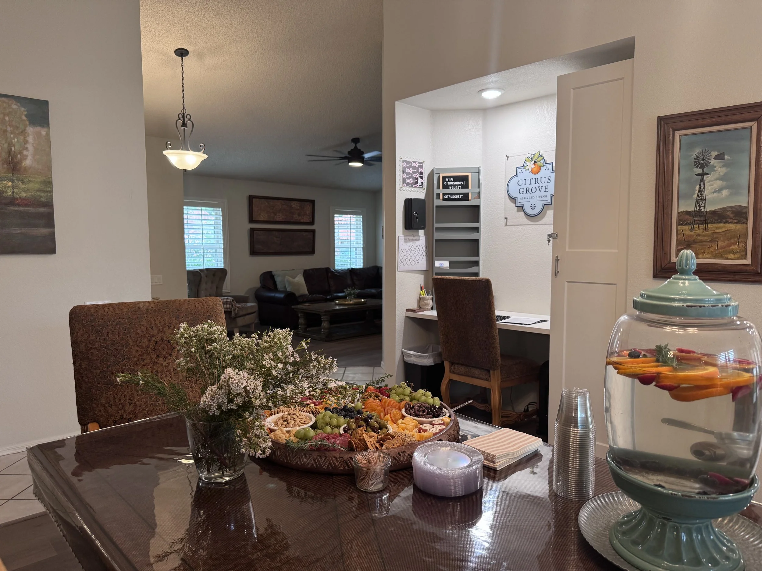 A dining room with a table filled with a large platter of assorted fruit and snacks, a bouquet of flowers, candles, and a tiered glass serving dish, with framed artworks on the walls, and a living area in the background with couches, windows, and ceiling fans.