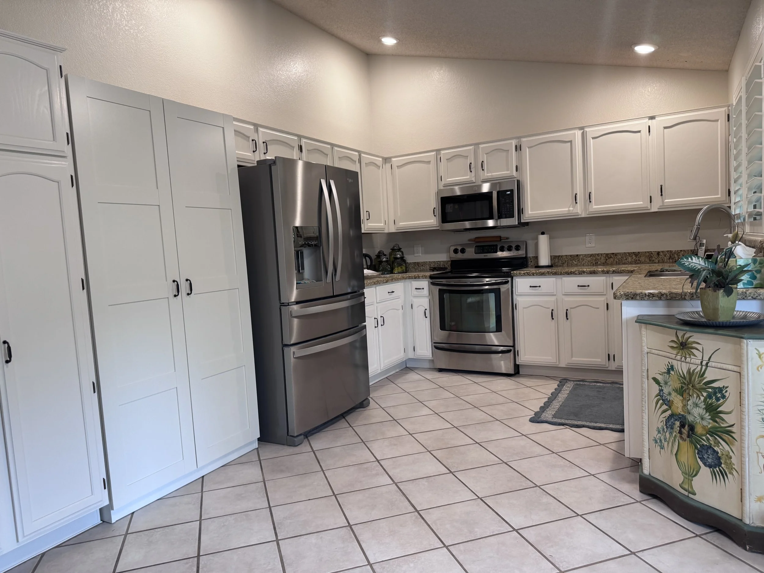 Kitchen with white cabinets, stainless steel refrigerator, microwave, oven, granite countertops, tiled floor, and decorative plants.