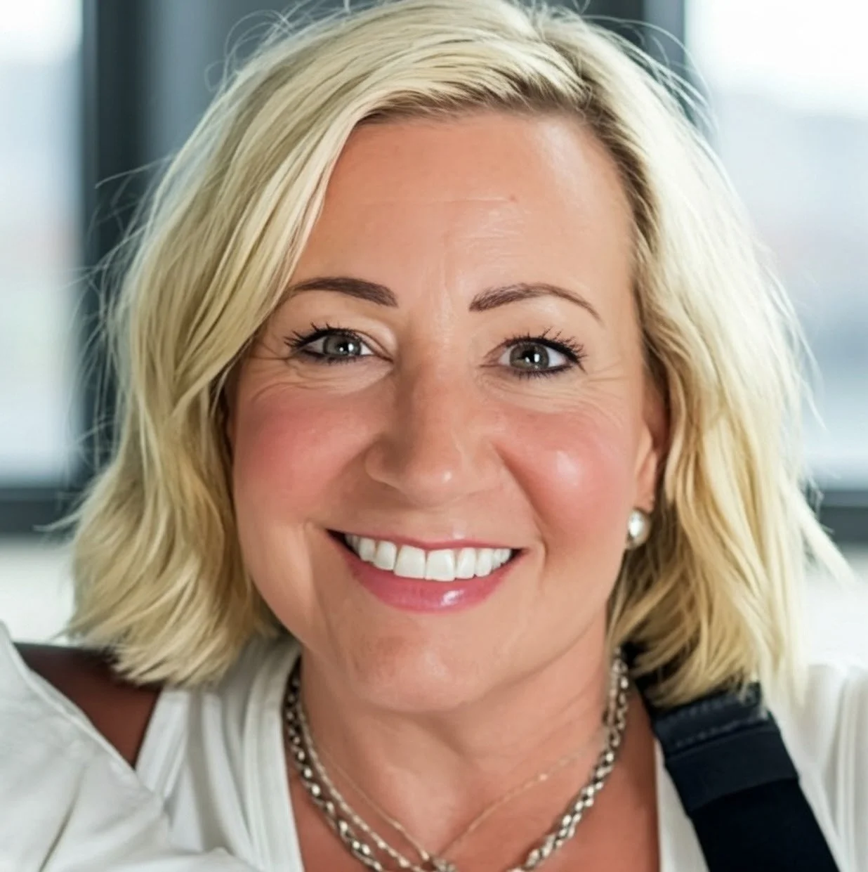 Close-up of a smiling woman with blonde hair, blue eyes, wearing pearl earrings, a silver necklace, and a white top.
