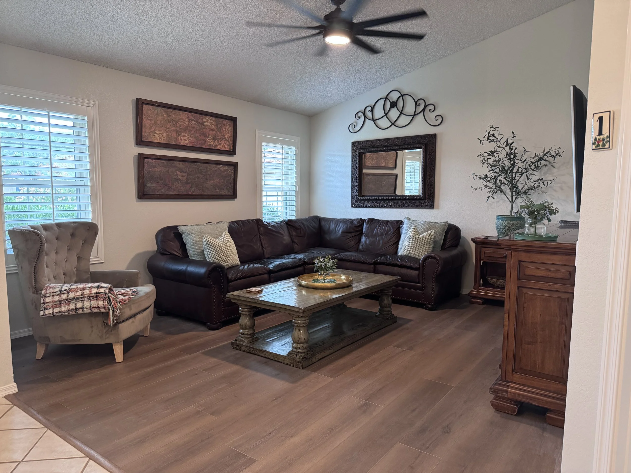 Living room with beige walls and wooden flooring. Contains a large brown leather sofa with white and beige cushions, a wooden coffee table with a decorative tray and flowers, a tufted beige armchair with a colorful blanket, two windows with white blinds, wall art, a mirror above the sofa, a ceiling fan, and a side table with a potted plant.