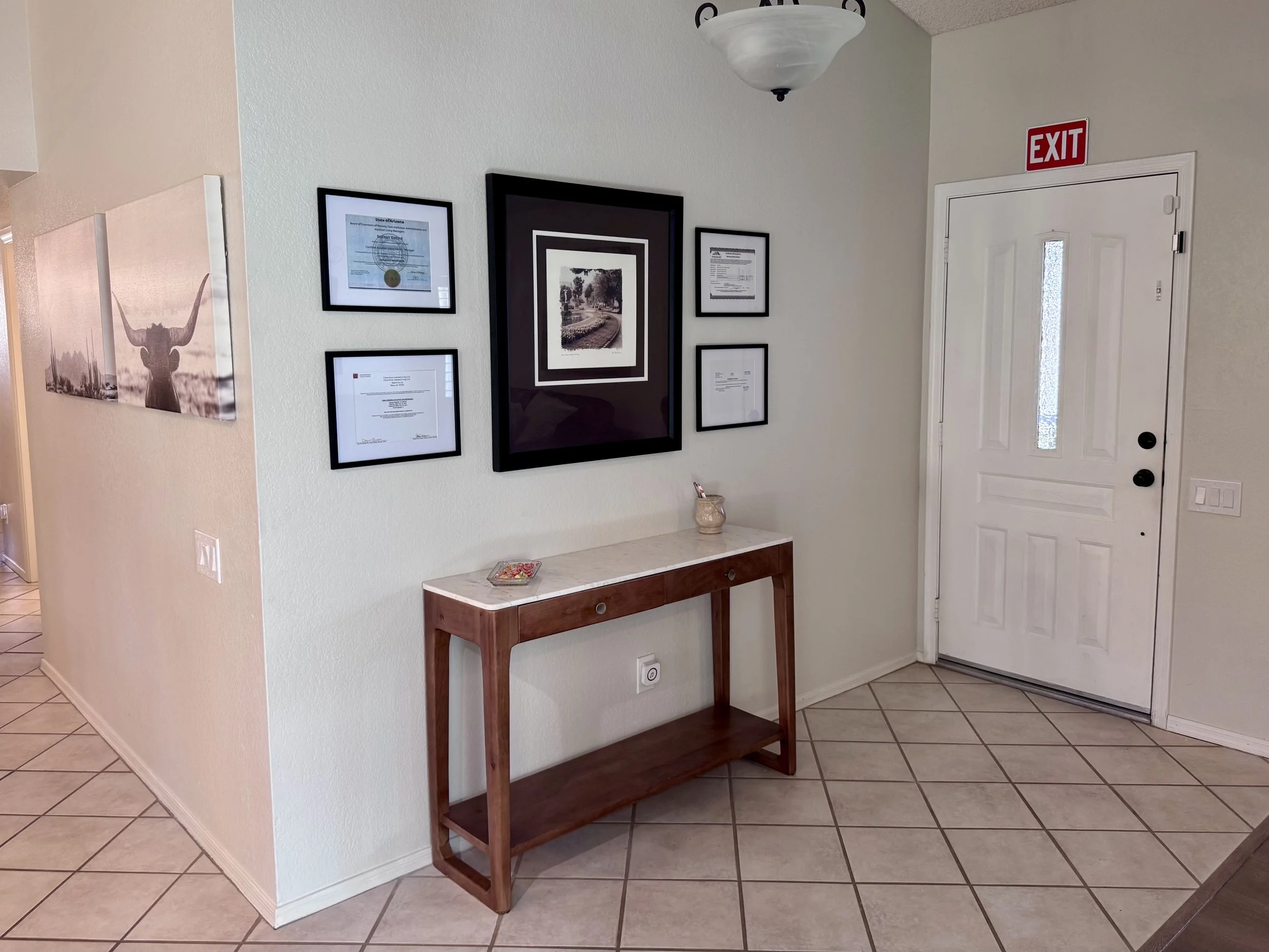 A hallway corner with framed certificates and artwork on the wall, a small wooden table with a bowl of candies, and a door with an illuminated glass panel and an exit sign above.