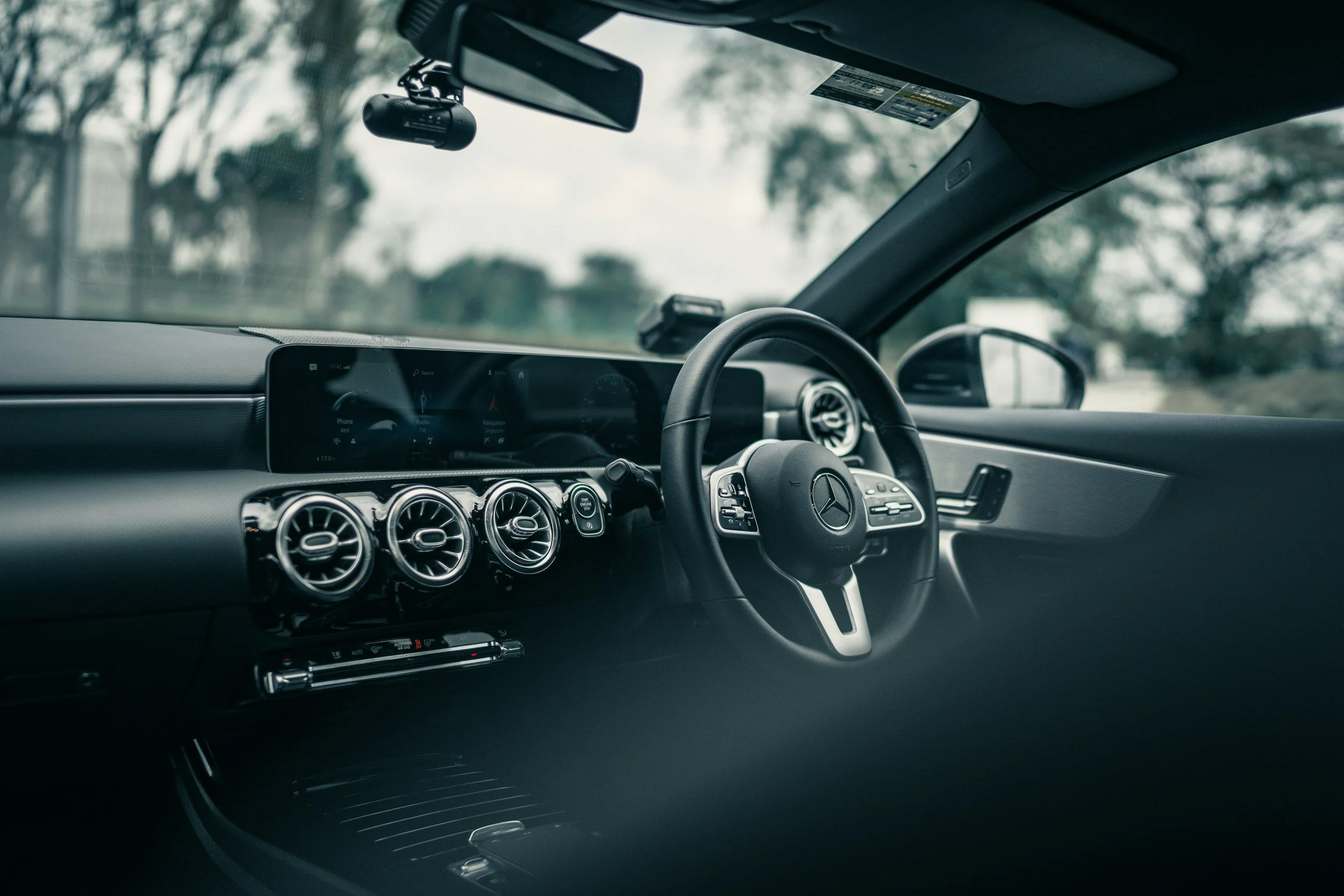 Interior of a luxurious black Mercedes-Benz car with chrome air vents, a digital infotainment system, and a steering wheel with the Mercedes-Benz logo. The car is parked outdoors with trees visible through the windshield.