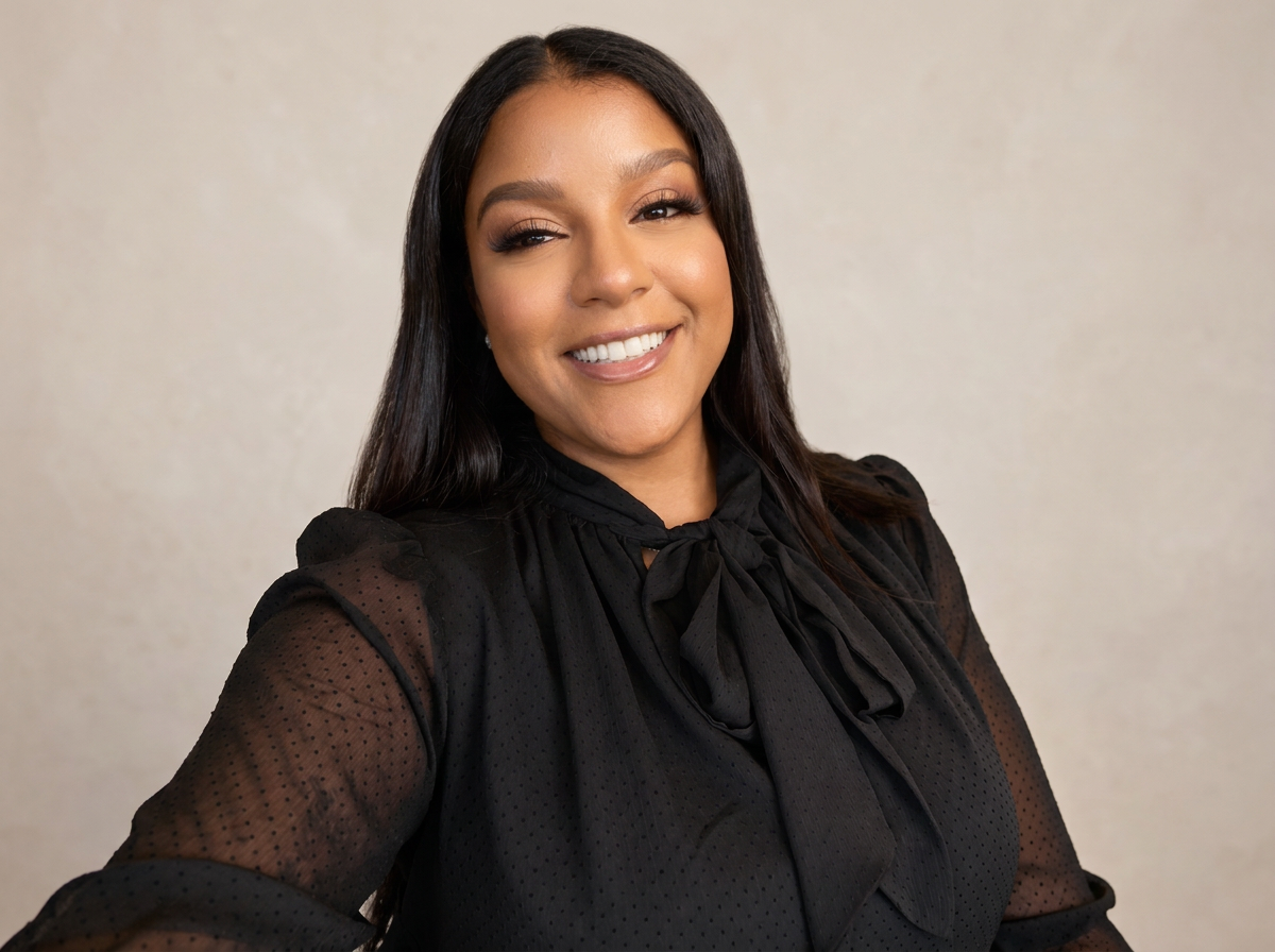 Smiling woman with long dark hair in a black blouse with a bow, standing against a neutral background.