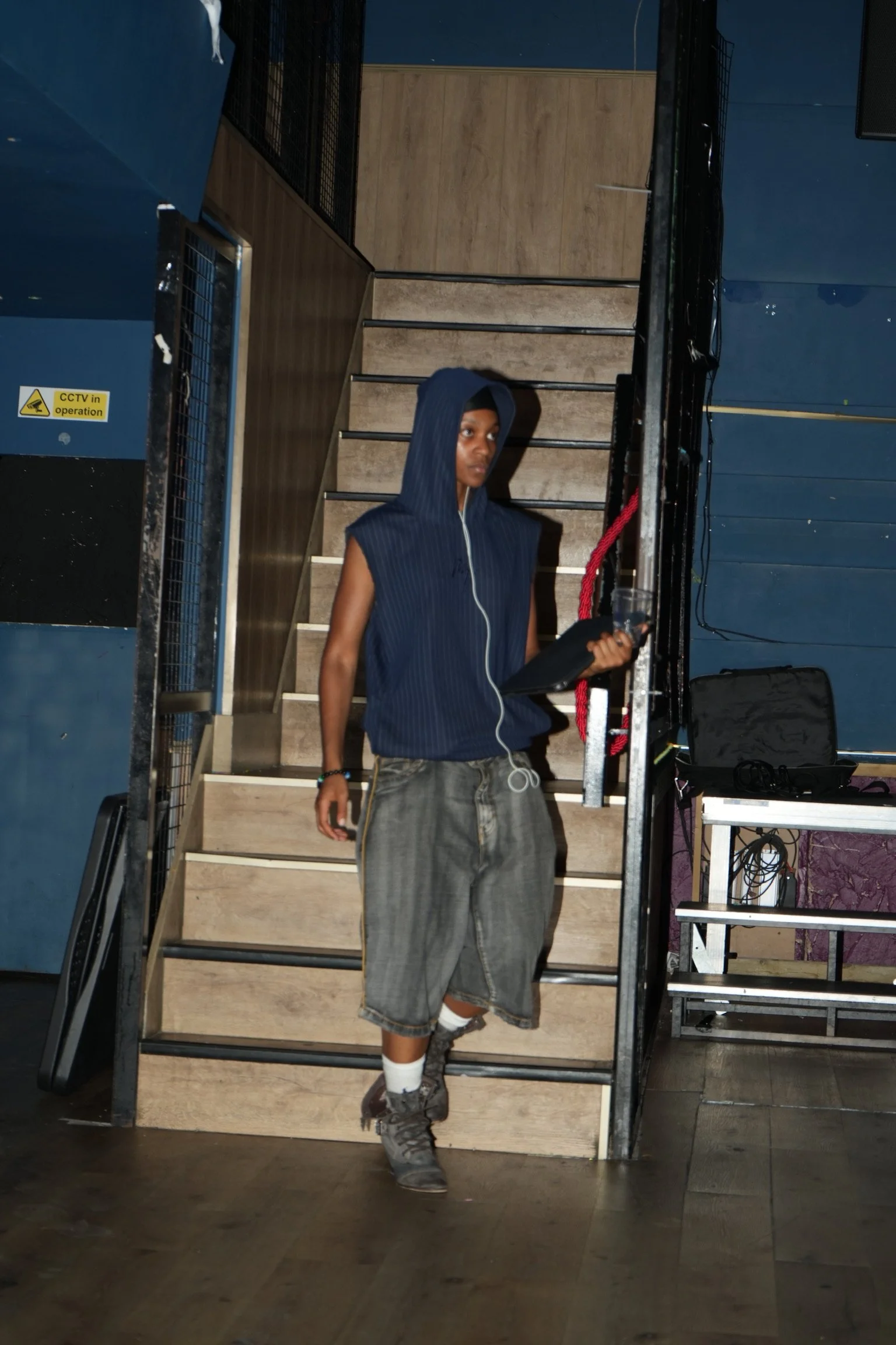 A young man with a hoodie, baggy jeans, sneakers, and earphones hanging from his pocket, standing on stairs inside a dark room or club. He is looking to his right.