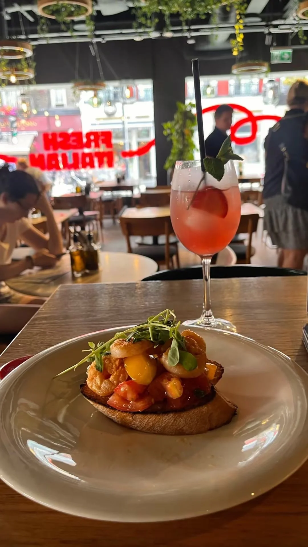 A plate of toast topped with shrimp, chopped tomatoes, mango, and microgreens. In the background, there is a pink cocktail garnished with a mint leaf and a straw, set on a wooden table inside a restaurant with large windows and many hanging plant decorations.