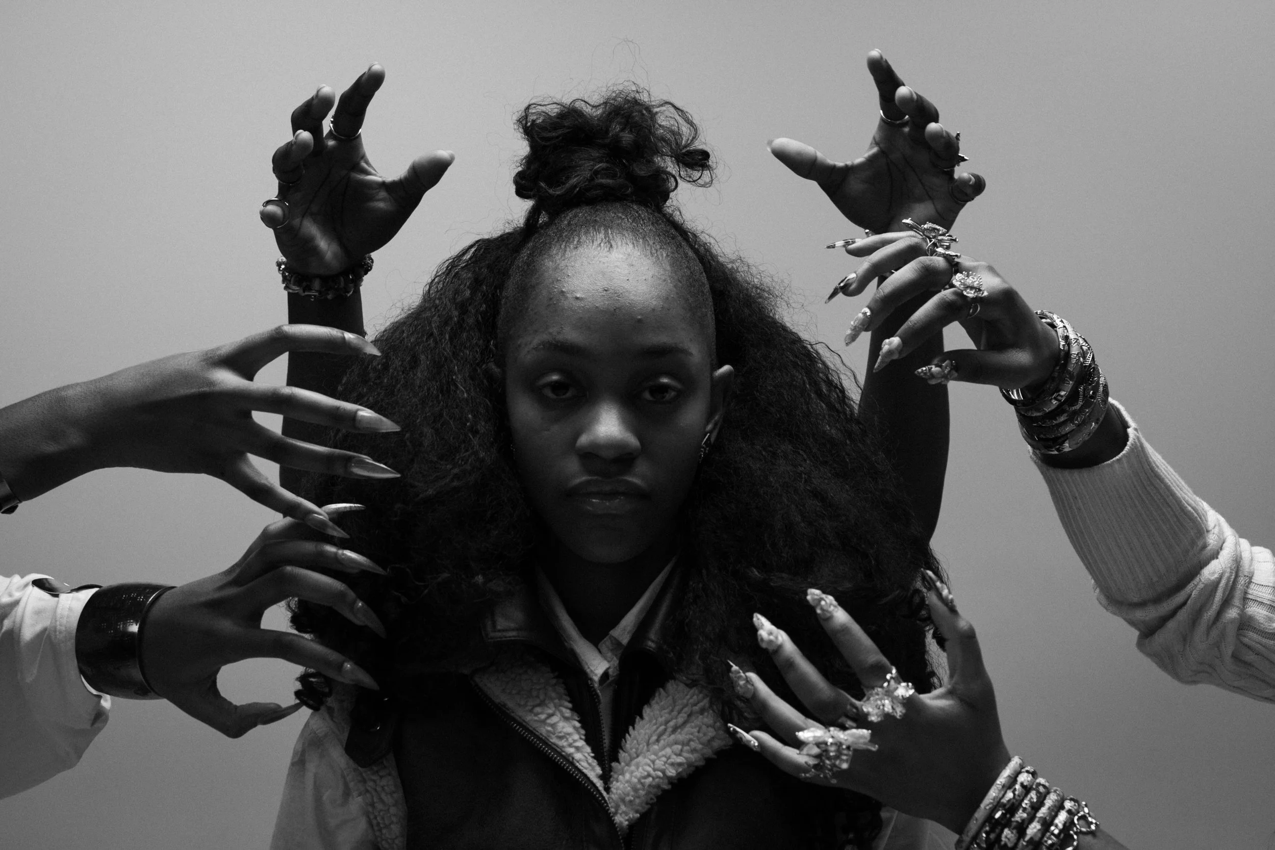 Black and white photograph of a young woman with curly hair, raising her hands and arms in artistic gestures, wearing multiple rings and bracelets.