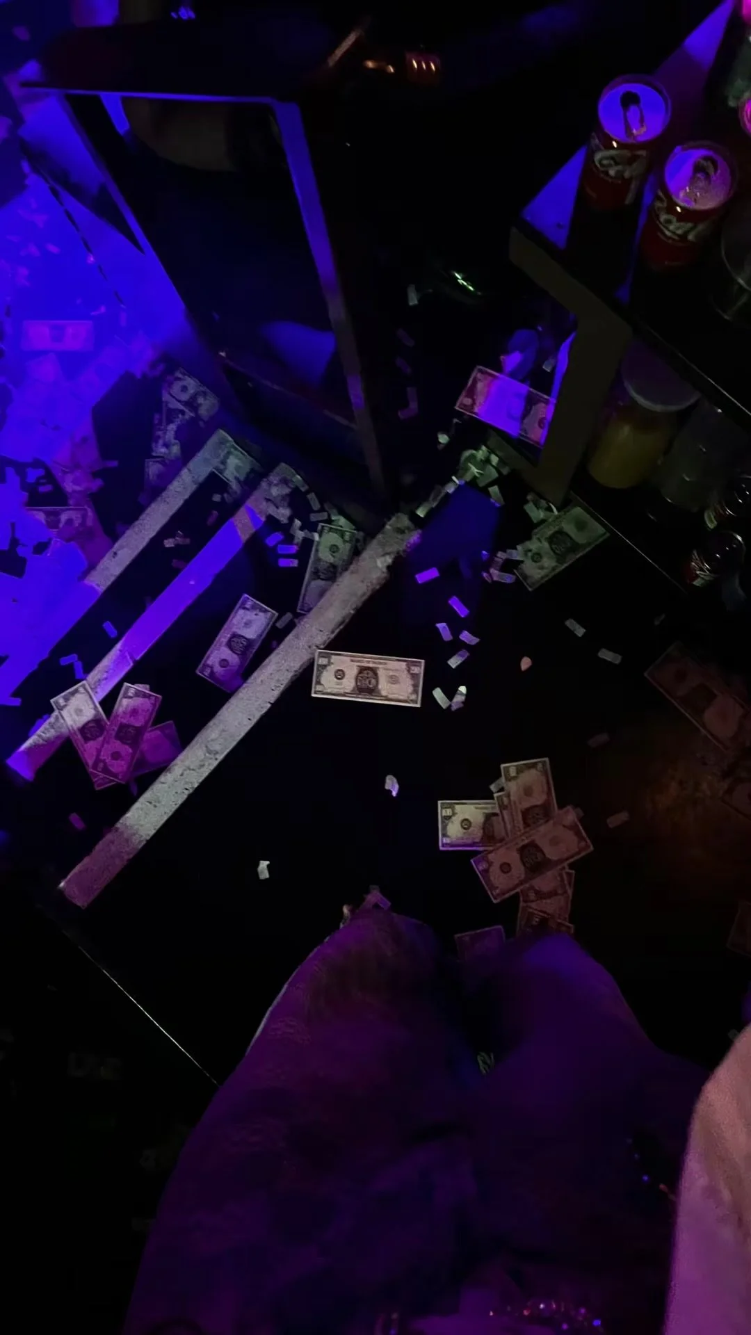Scattered dollar bills and confetti on the floor of a dark room with purple lighting, part of a person's leg and shoe visible in the foreground.