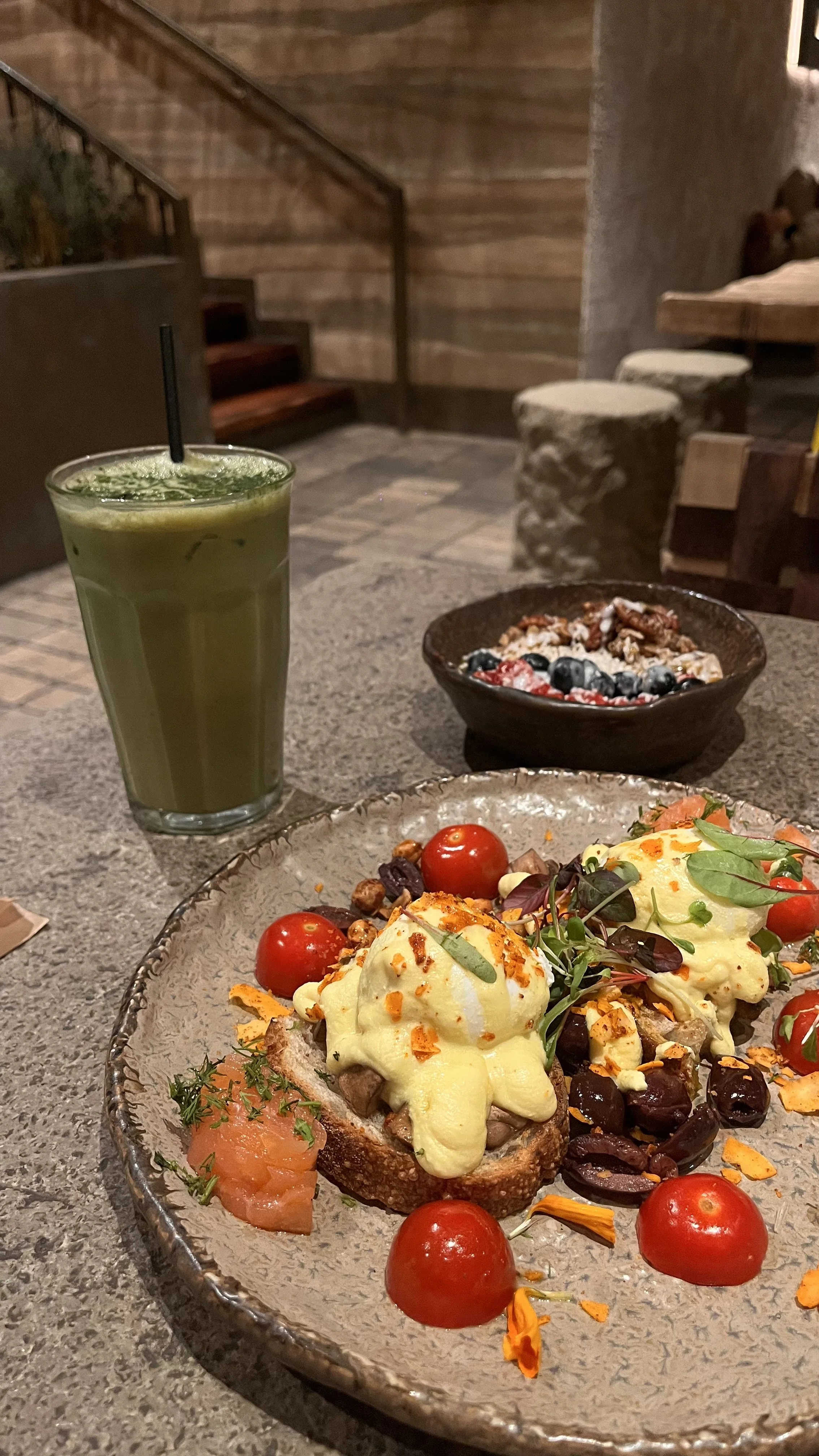 A meal on a rustic table with a dish of eggs Benedict topped with microgreens, cherry tomatoes, and orange peel, a small smoked salmon with herbs, a bowl of mixed berries with granola, and a tall glass of green juice.